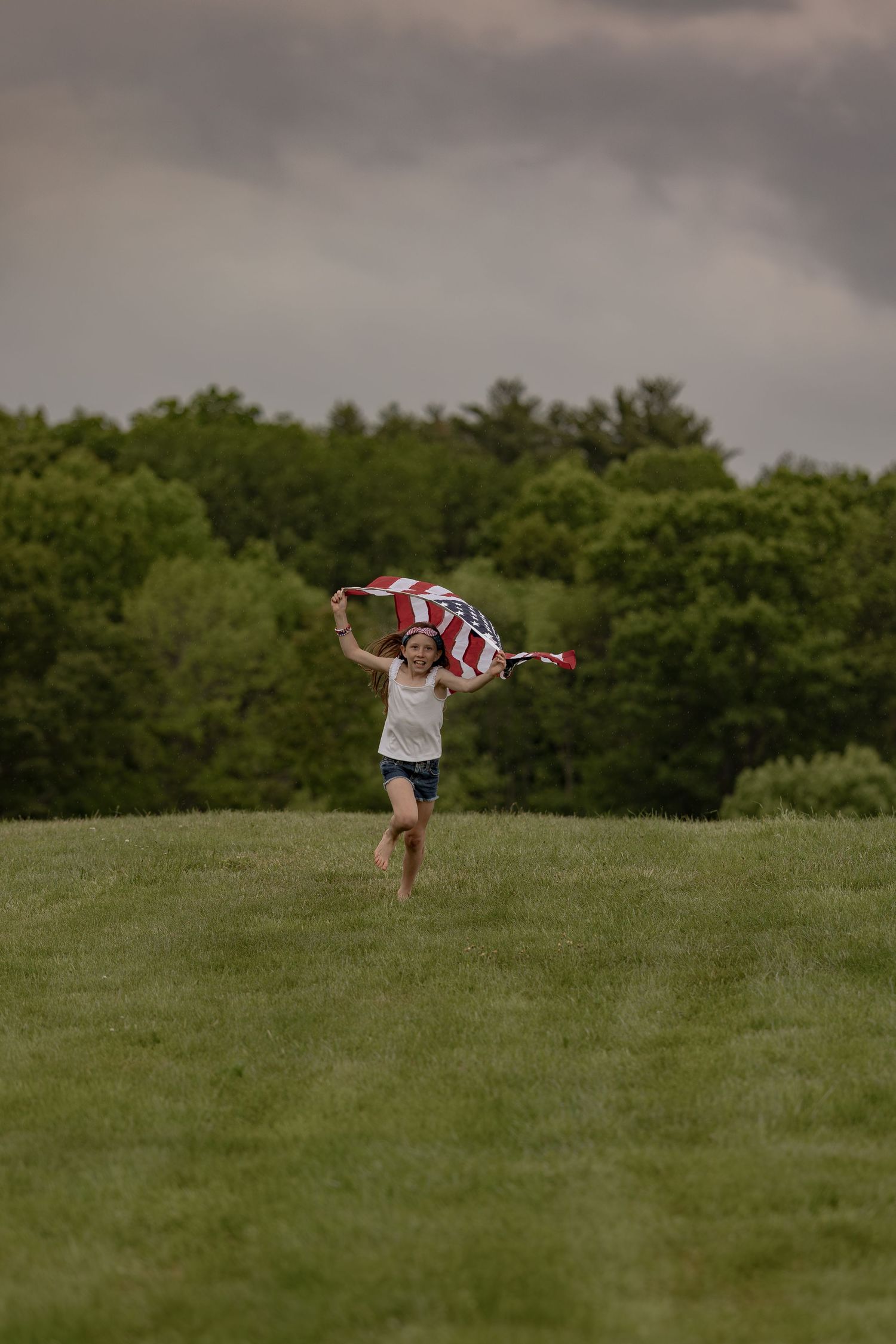Young girl running through green field holding American flag overhead under dramatic cloudy sky.