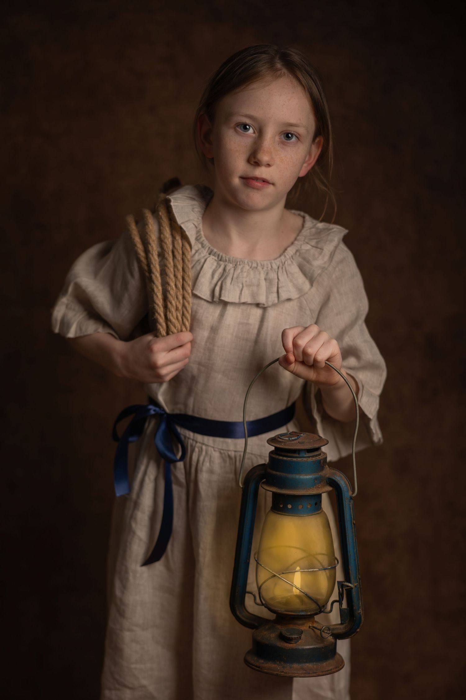 Young girl in vintage linen dress holding a glowing blue kerosene lantern and rope bundle against dark background.