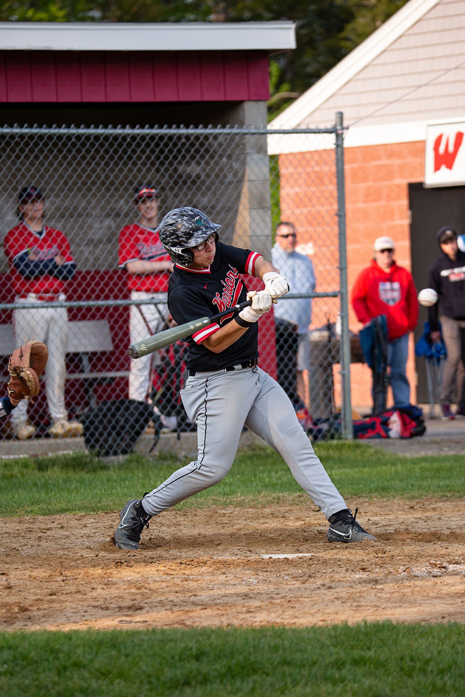 Baseball batter in black and gray uniform swings at pitch during a game, with dugout and spectators visible in background.