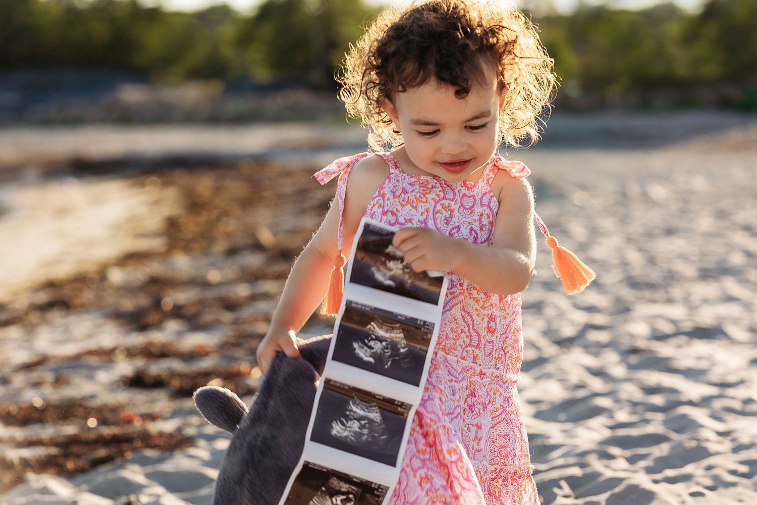 A person in a floral dress holds an ultrasound photo while standing on a sandy beach at sunset.