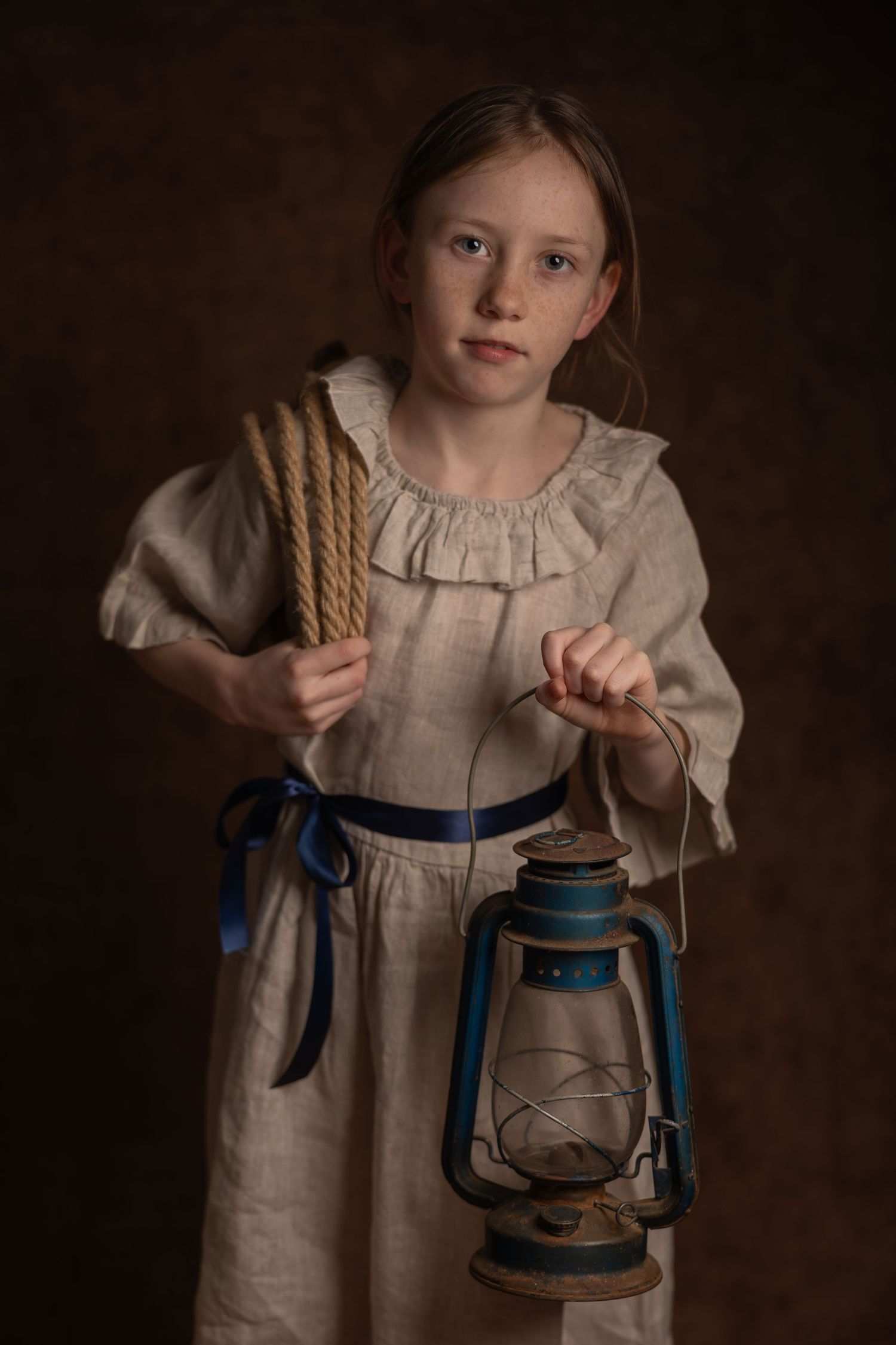Young girl in linen dress holding vintage blue oil lantern and rope bundle against dark background.