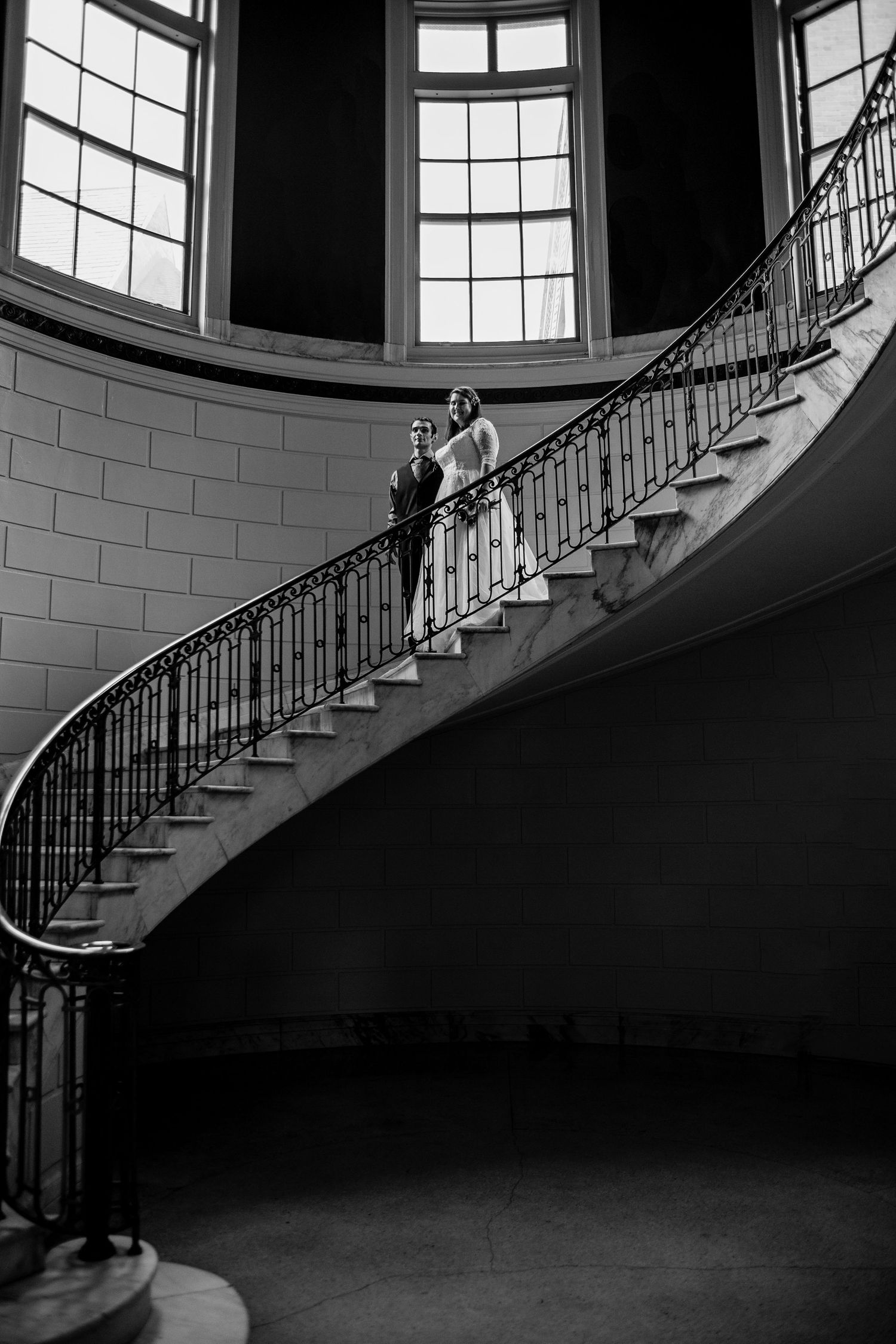 Black and white photo of a bride and groom posing on an elegant curved staircase with ornate iron railings.