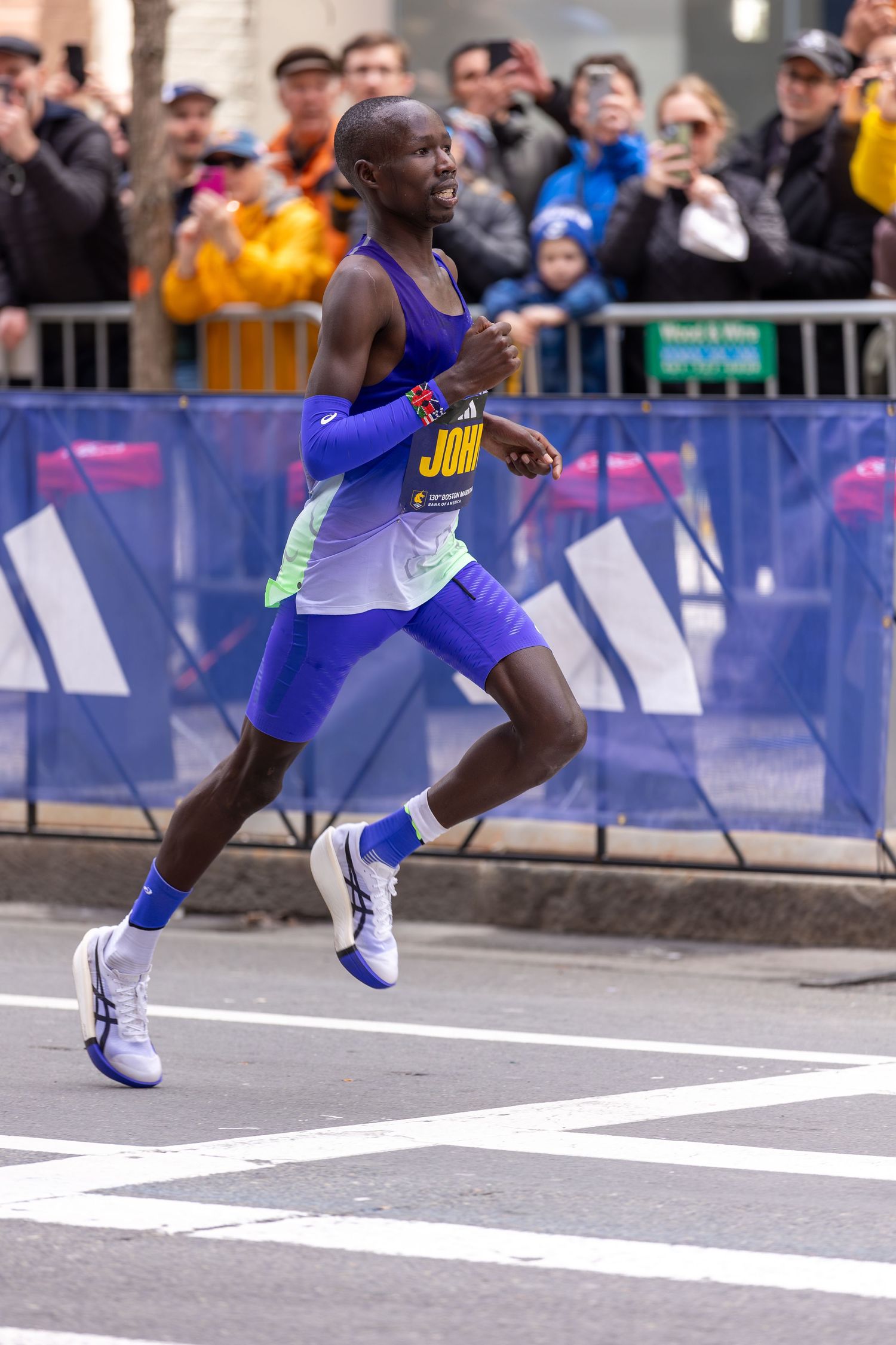 Male marathon runner in purple Adidas kit racing past spectators at a major city marathon event.