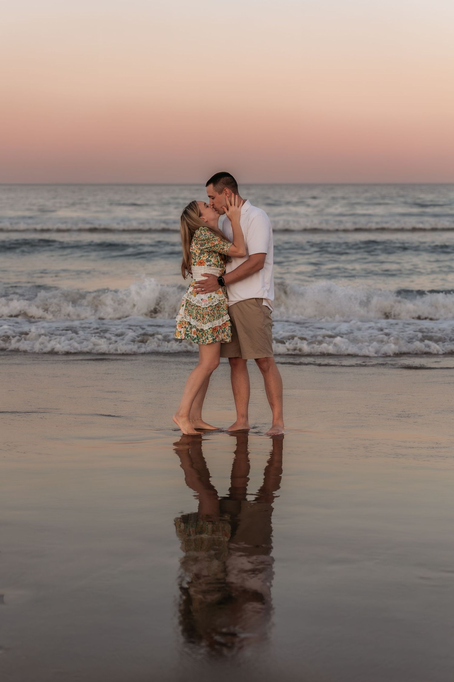 Couple sharing a romantic kiss on a wet beach at sunset, with ocean waves and pink sky reflected on the sand.