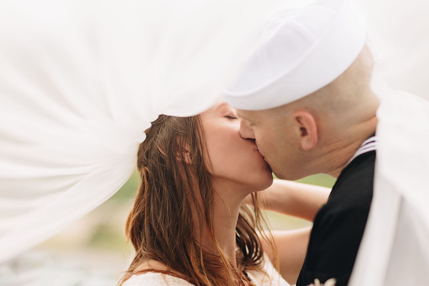 A bride and groom share a romantic kiss under a flowing white wedding veil outdoors on their wedding day.
