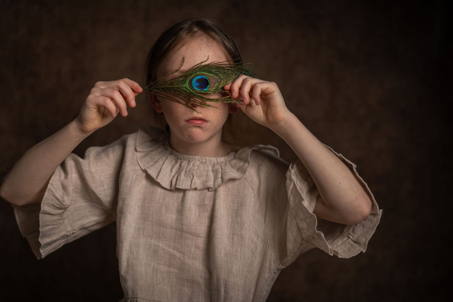 A young girl in a linen dress holds a peacock feather over her eyes against a dark background.
