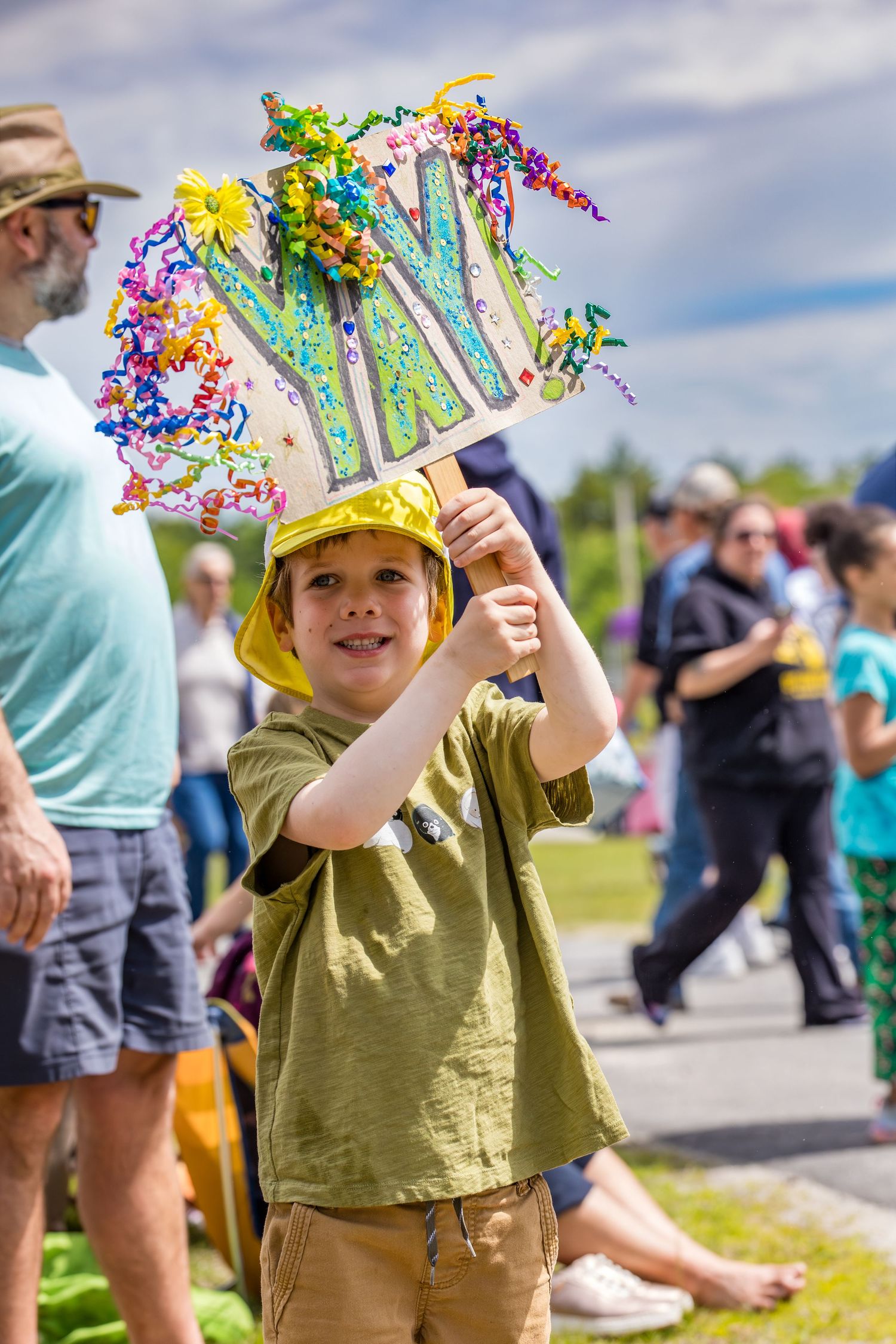 Young boy smiling at outdoor festival, holding colorful decorated hat with streamers and ornaments at a community parade.