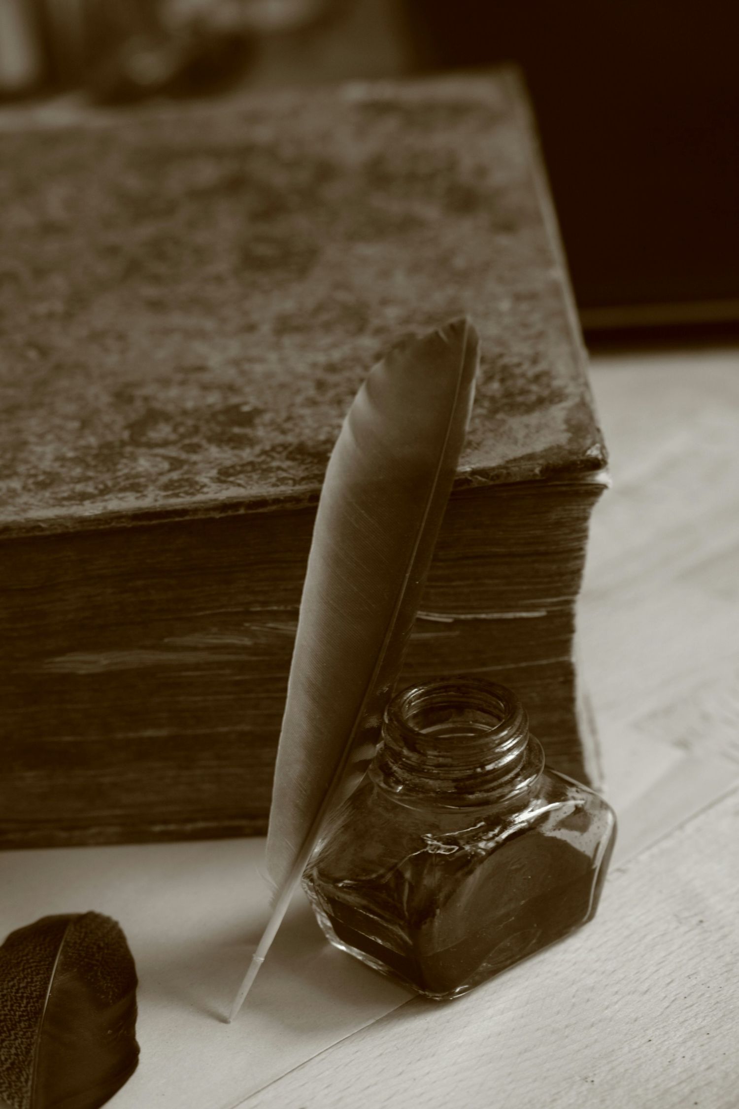 A vintage sepia photograph shows a quill pen and ink well on a rustic wooden table with parchment paper.