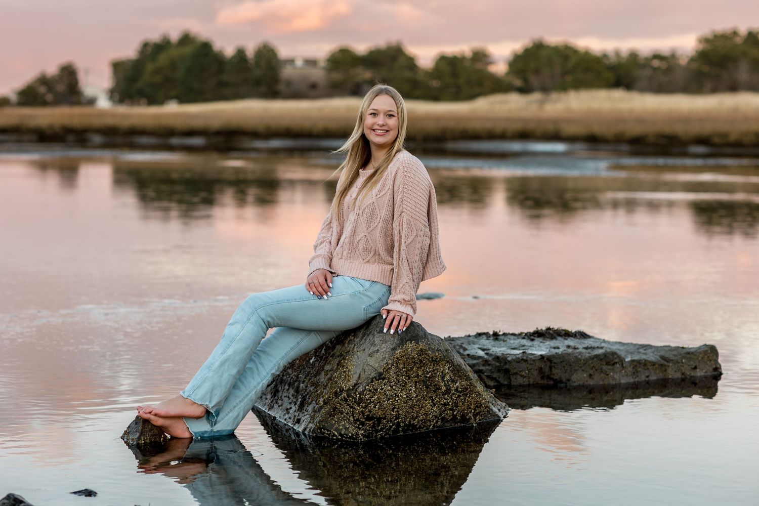 Young woman in casual attire sitting on a log by a serene lake during sunset.