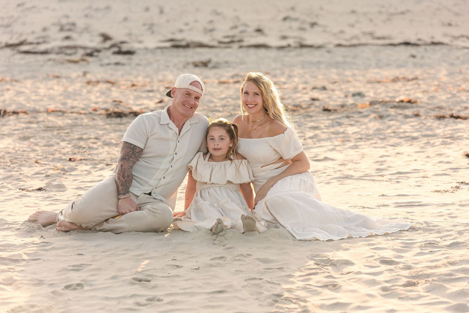 Family dressed in white sitting together on sandy beach at sunset.