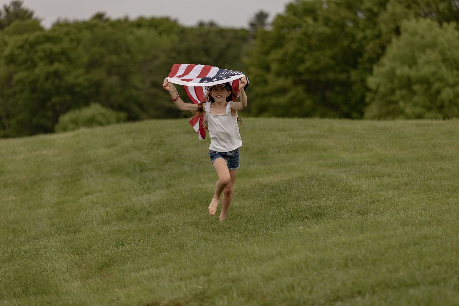 Young girl running barefoot on green grass while holding an American flag overhead on a cloudy day.