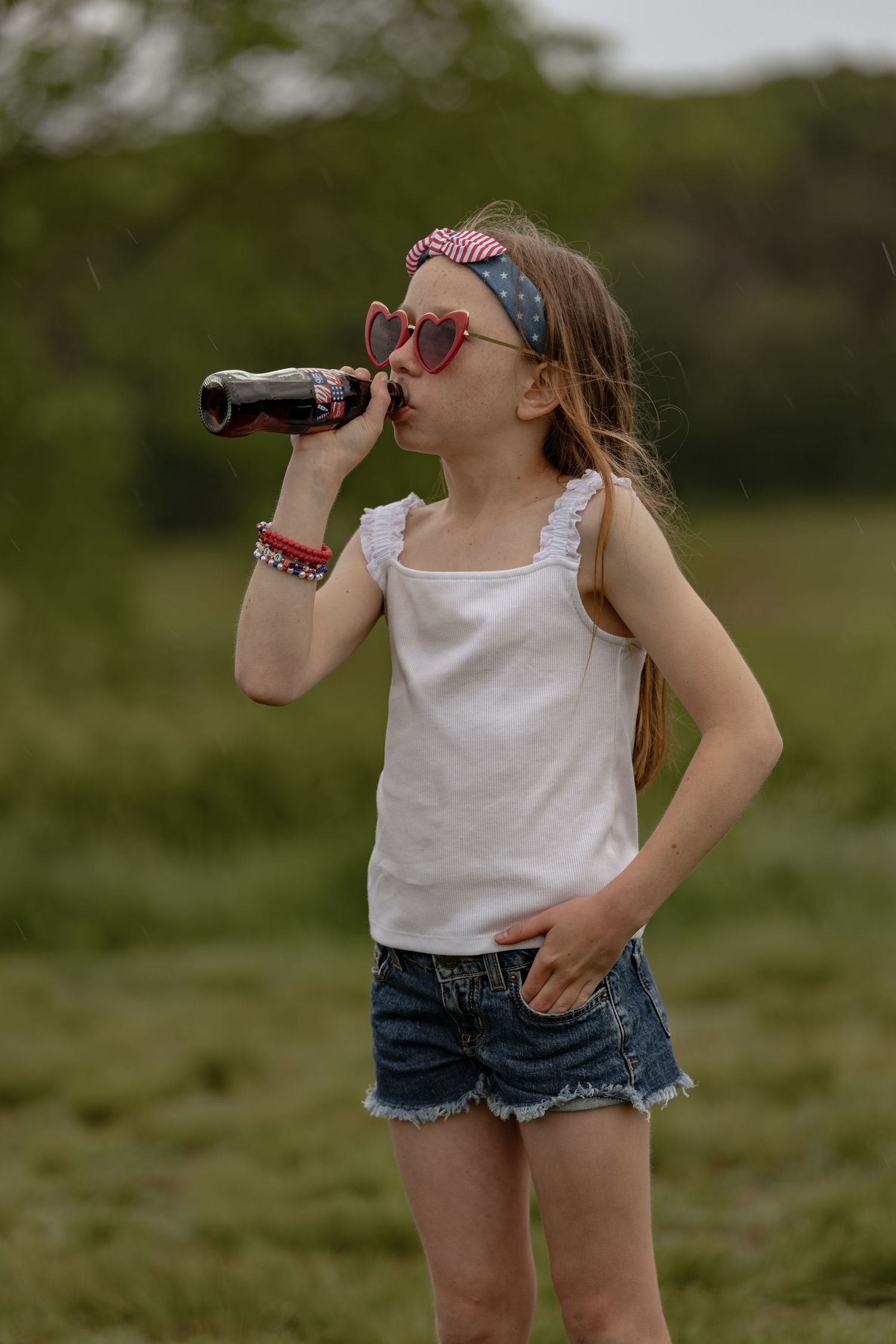 Young girl in white tank top and denim shorts drinking soda bottle outdoors wearing heart sunglasses and patriotic headband.