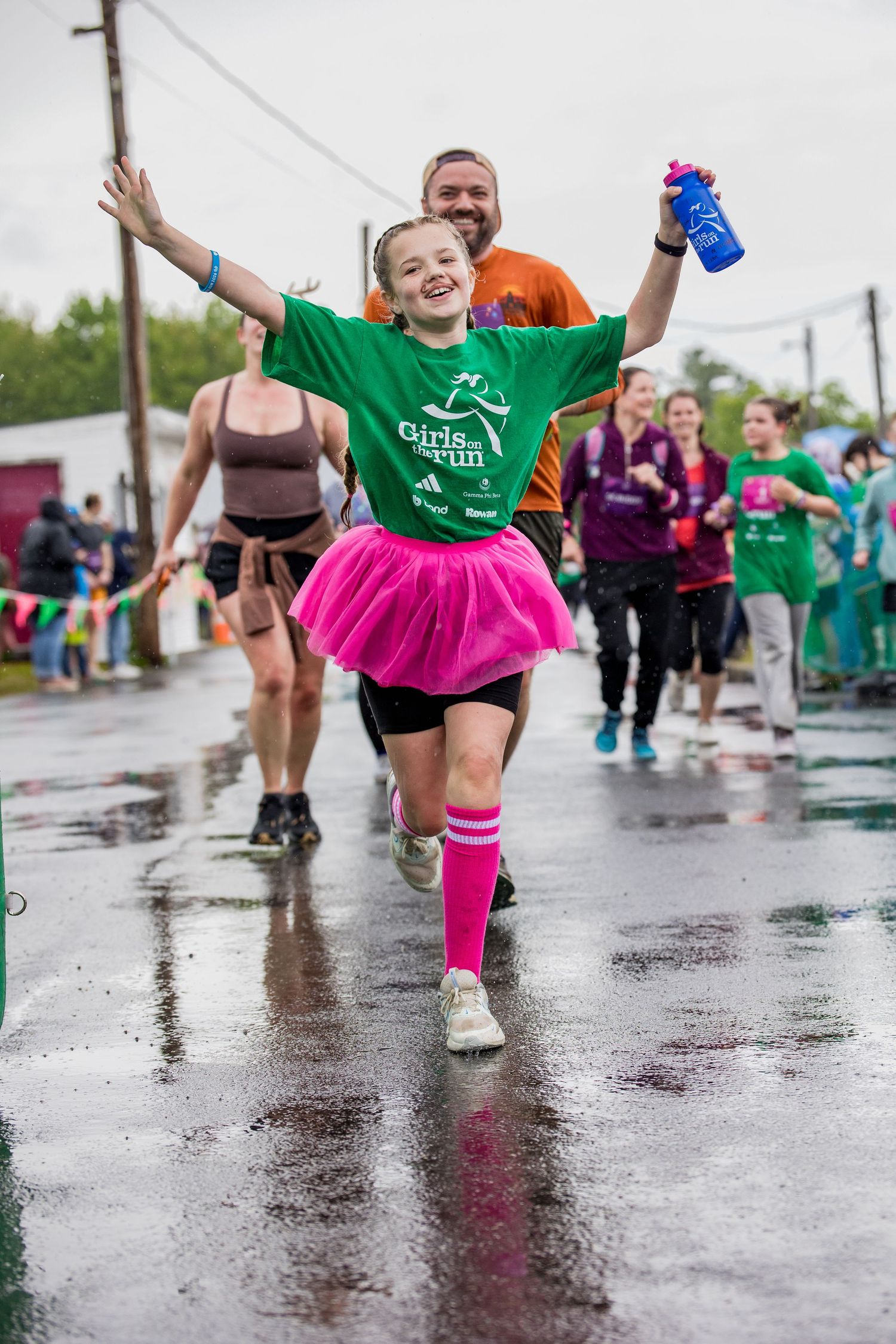 Joyful girl in green Girls on the Run shirt and pink tutu celebrates finishing a rainy 5K race with arms raised.