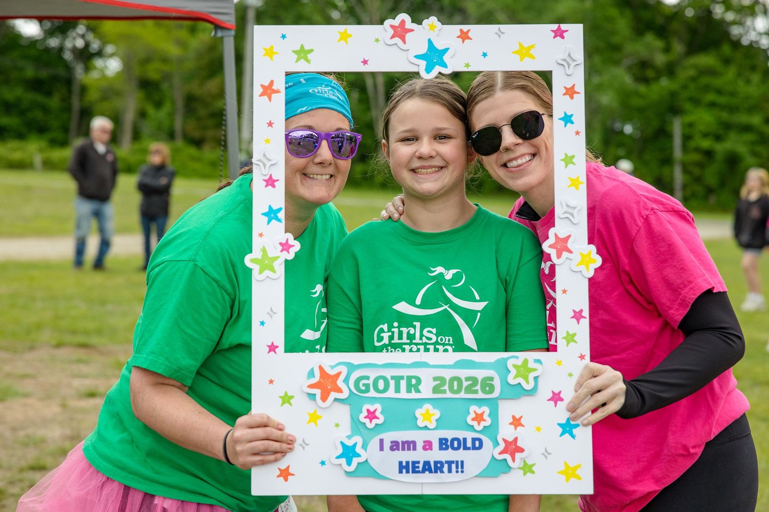 Three smiling girls pose with a GOTR 2026 Bold Heart photo frame at a Girls on the Run outdoor event.