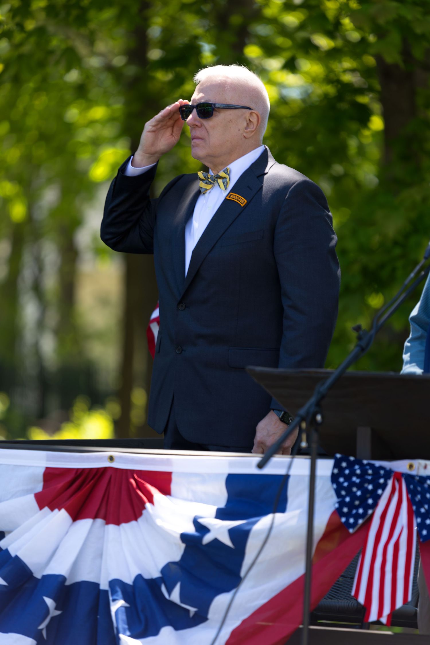 Elderly man in suit and bow tie saluting at patriotic American flag ceremony outdoors.