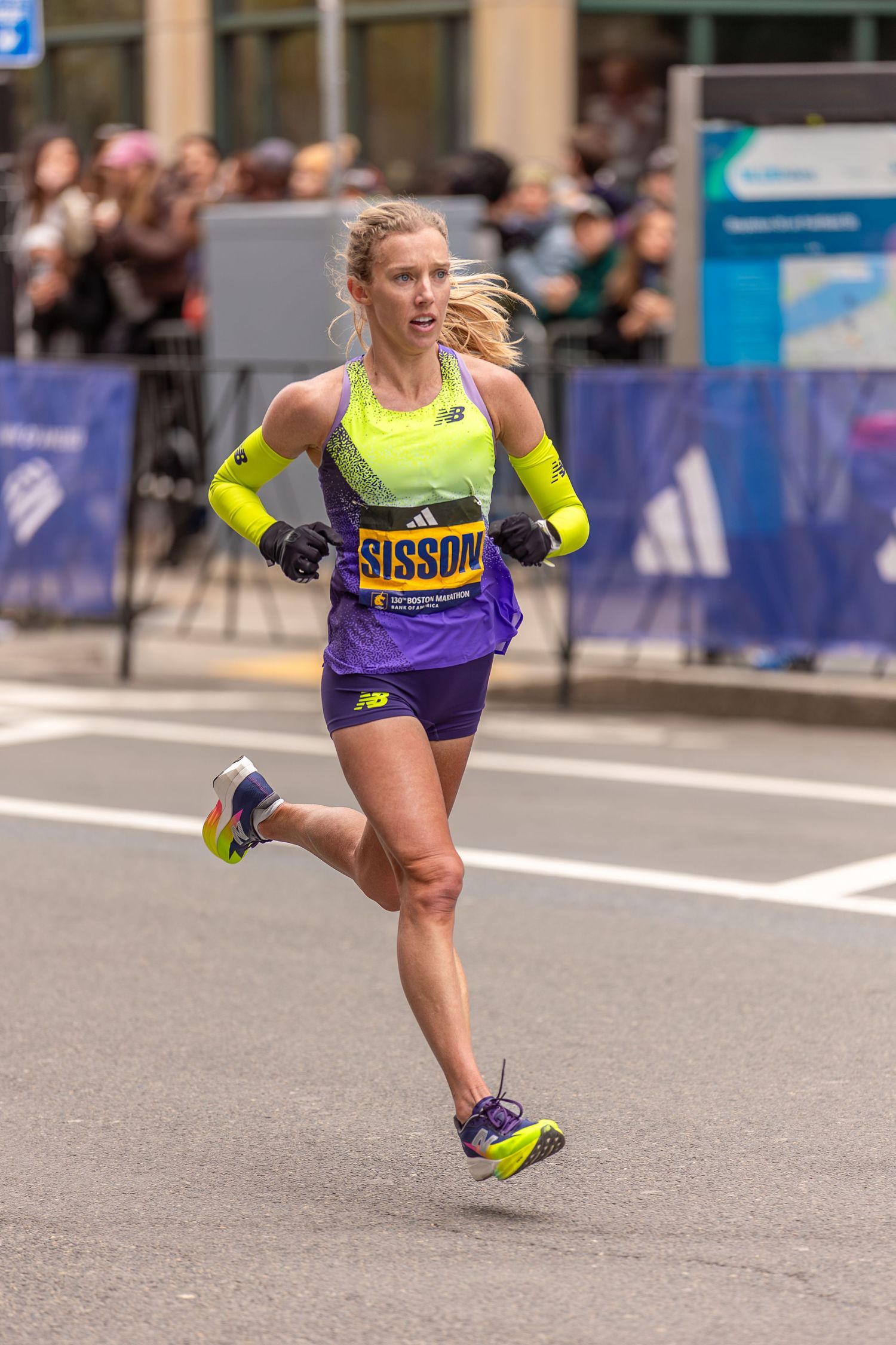 Runner Sisson competing in a marathon wearing New Balance neon yellow and purple athletic gear on a city street.