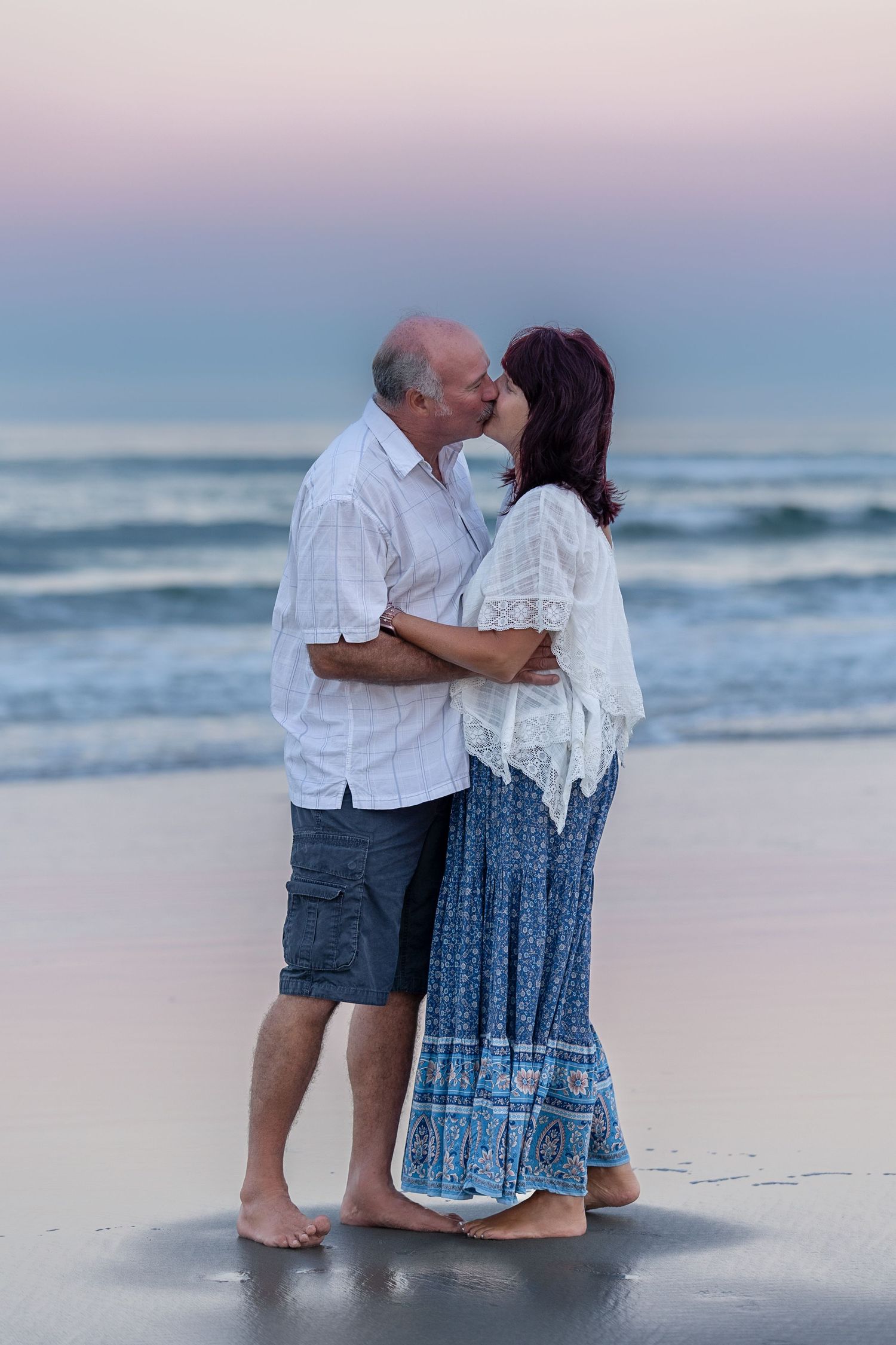Elderly couple sharing a romantic kiss on a sandy beach at sunset, waves and pastel sky in the background.