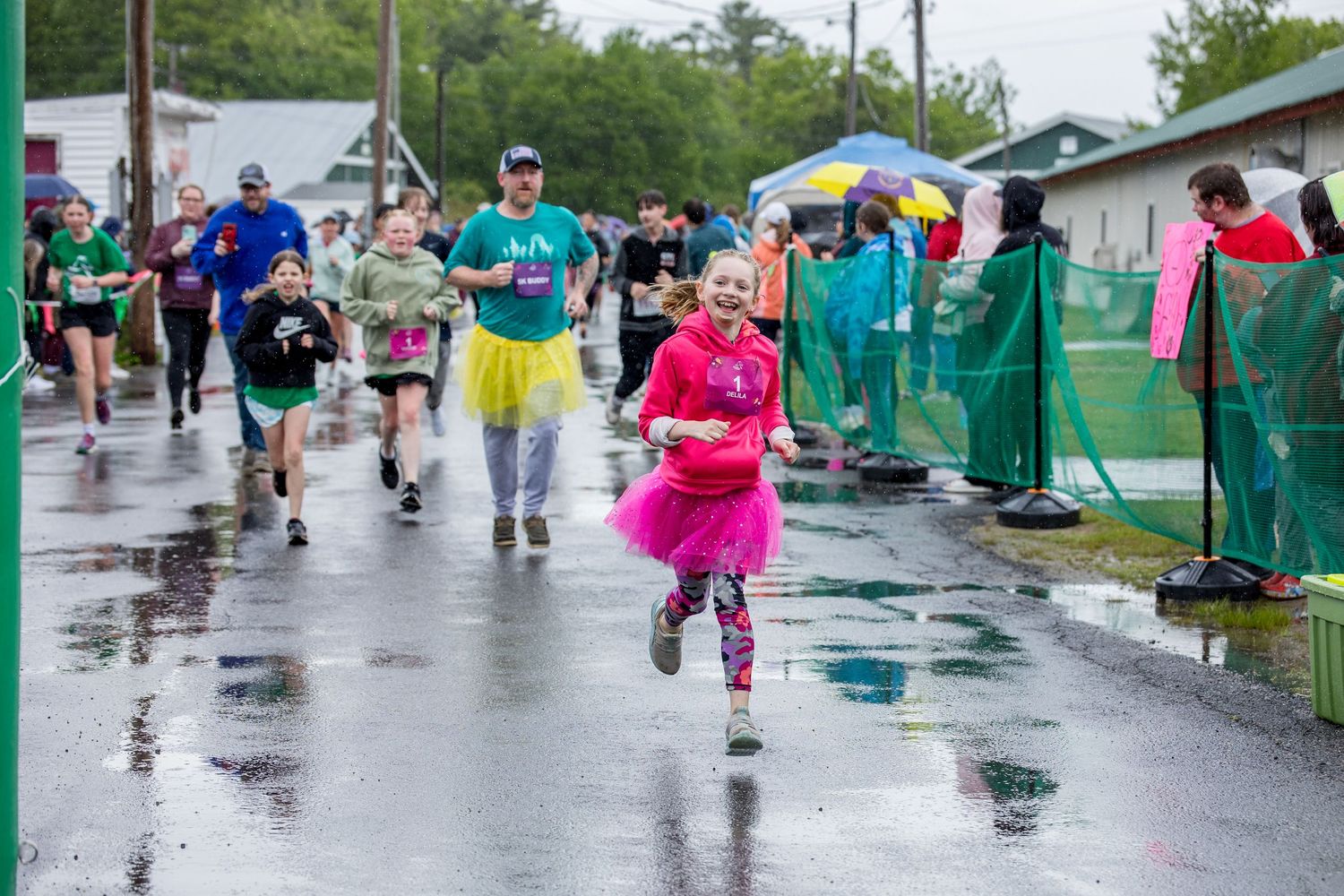 Children and adults race in colorful tutus during a fun run event on a rainy day, splashing through puddles.