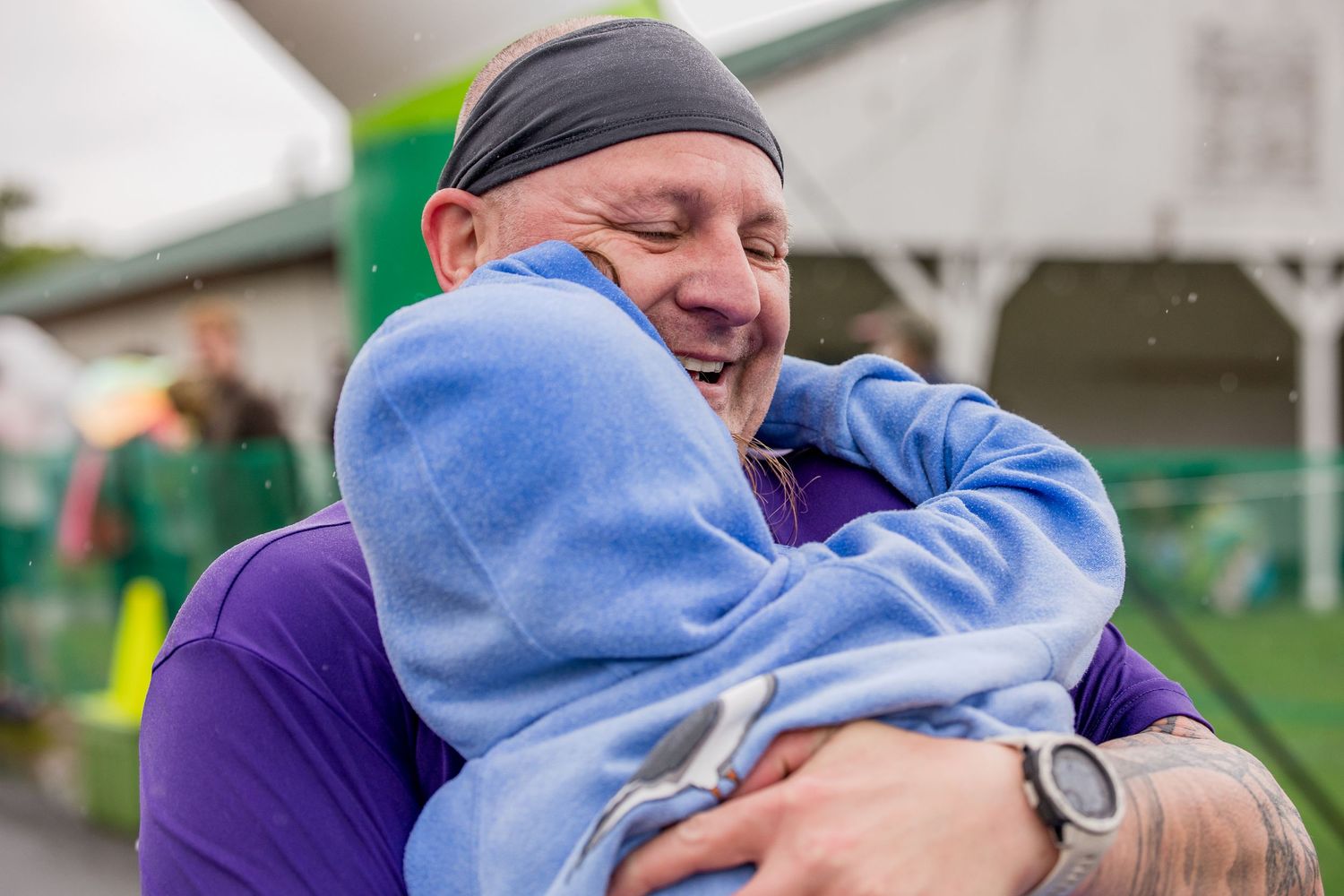 A bald man in a purple shirt smiles while sharing a warm embrace with someone in a blue fleece jacket outdoors.