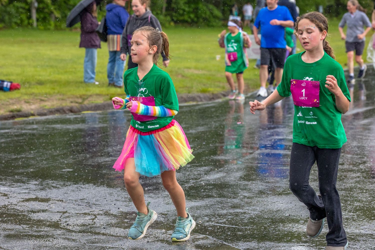 Two girls in green shirts running a race outdoors, one wearing a colorful rainbow tutu skirt on a wet pavement.