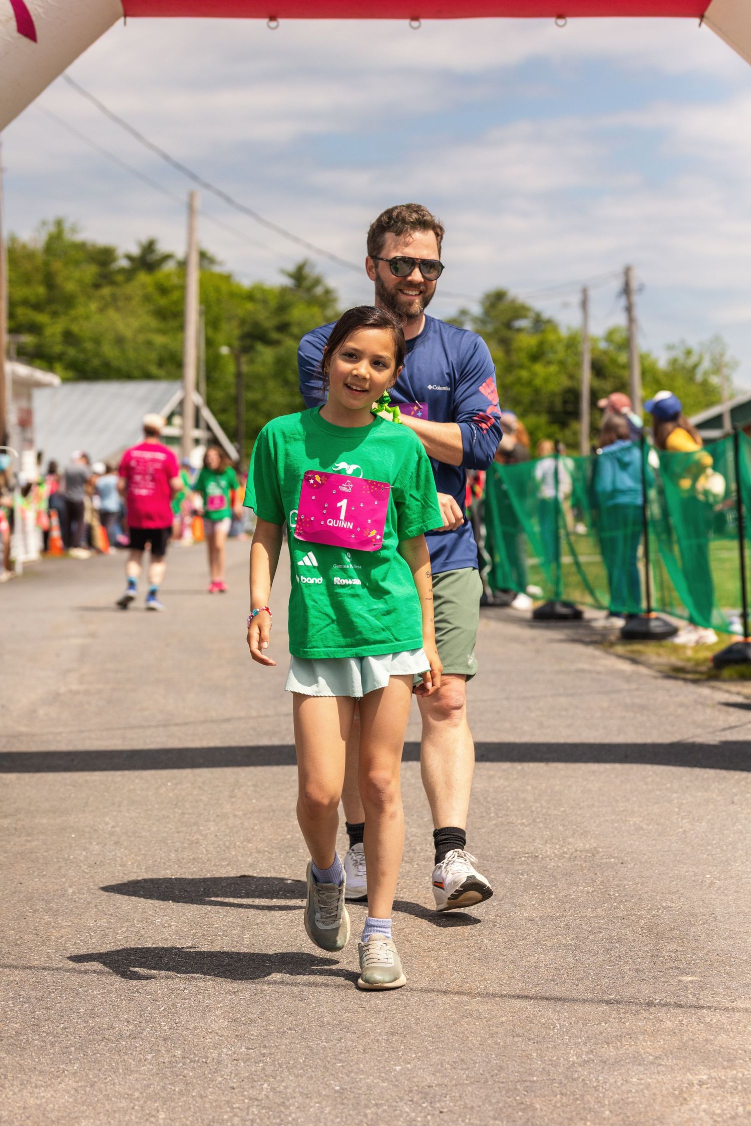 A girl in a green shirt with a pink race bib crosses the finish line at a fun run, guided by a smiling man.