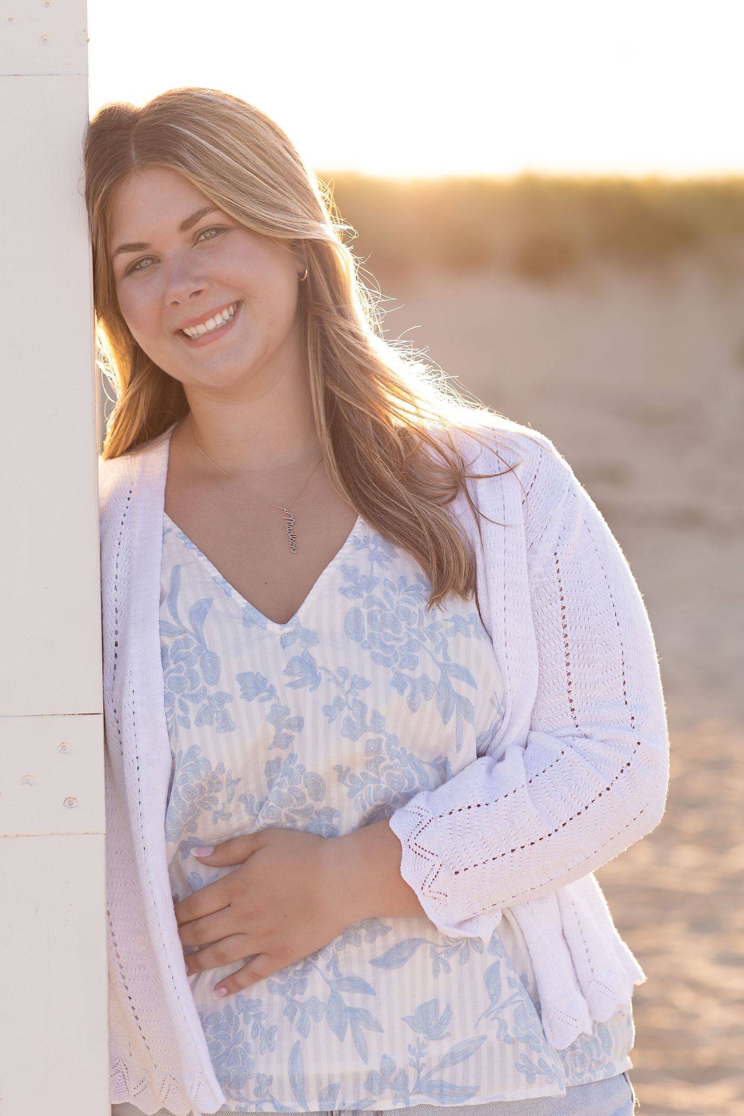 A graduating senior in a white lace cardigan and blue tie-dye top poses at sunset on the beach.