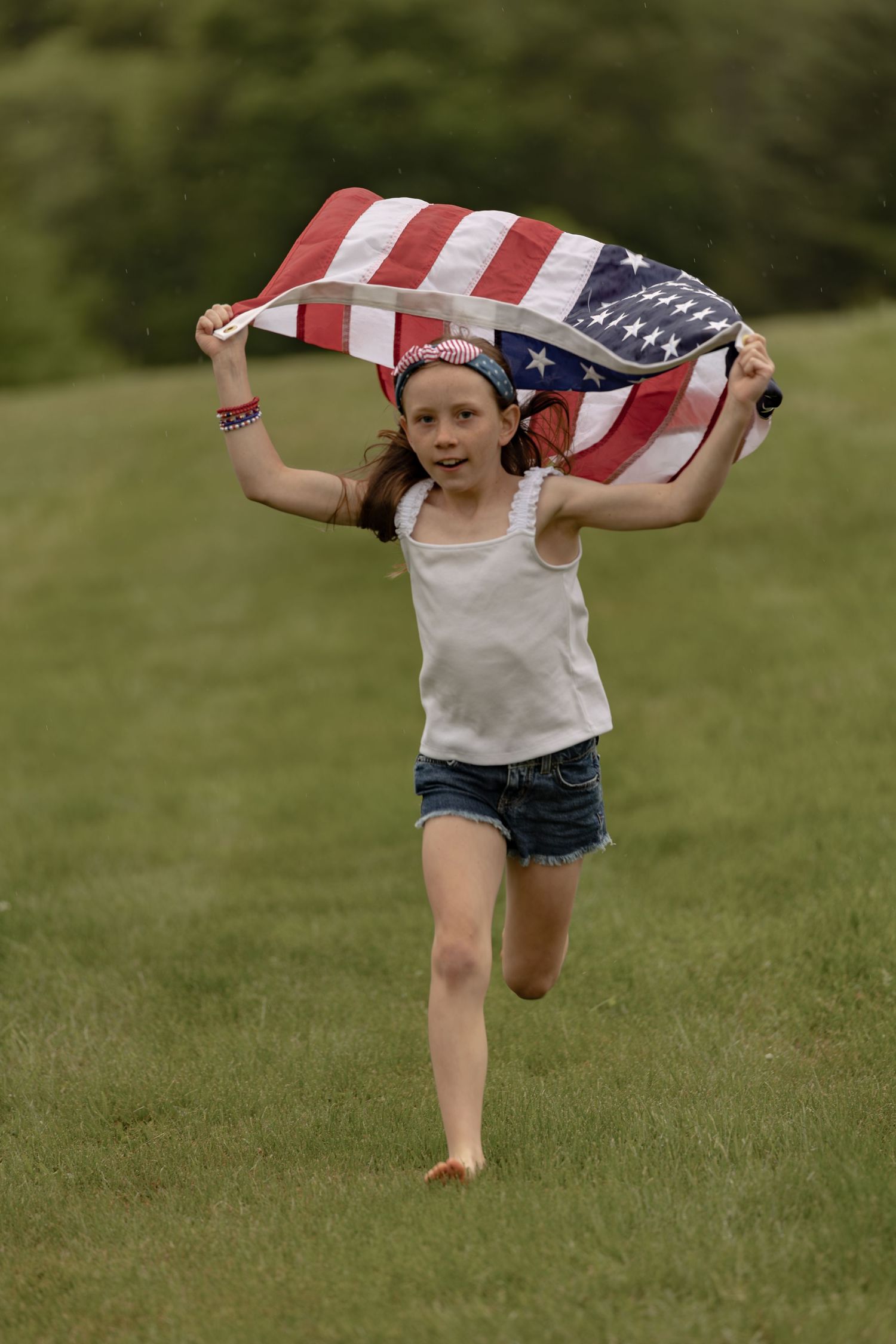 A young girl runs across a green field holding an American flag above her head, wearing denim shorts and a patriotic bandana.