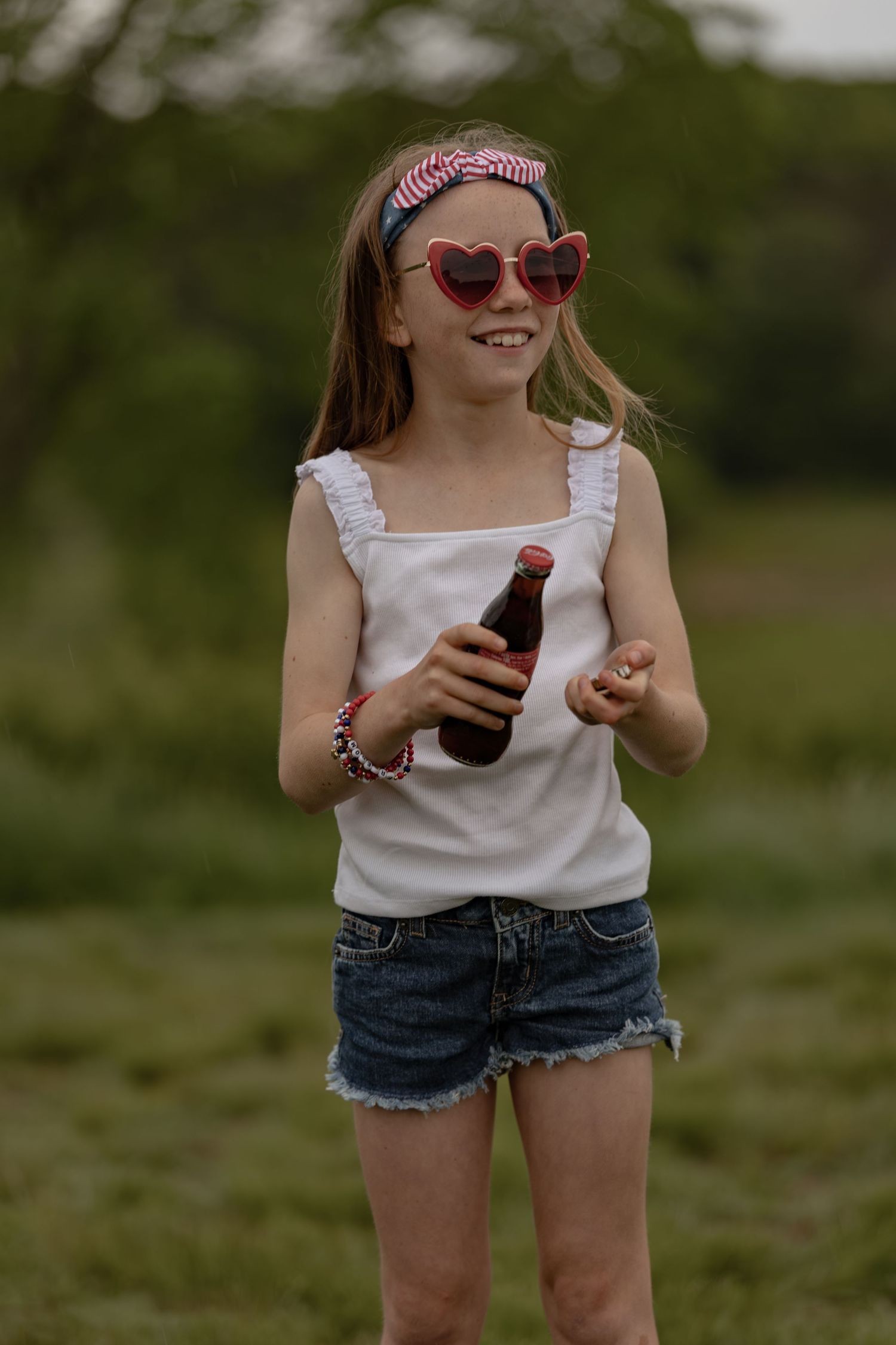 Young girl in white tank top and denim shorts wearing heart-shaped sunglasses, holding a soda bottle outdoors.