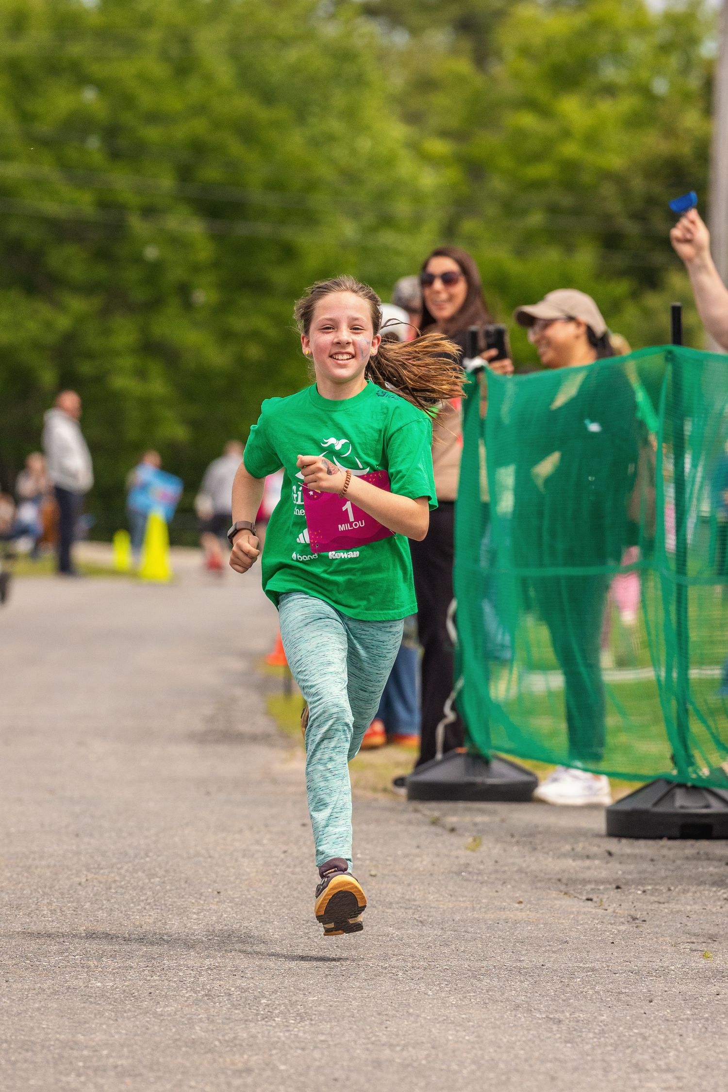 Young girl in green t-shirt smiling while running a race on a paved path with spectators cheering.