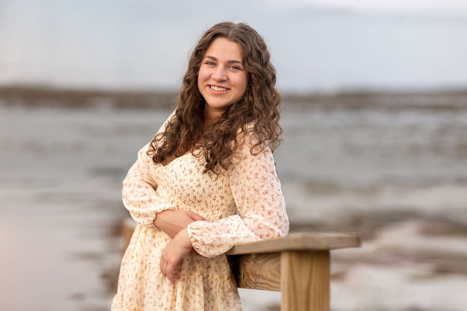 Someone in a cream polka dot dress poses by a wooden railing at the beach on an overcast day.