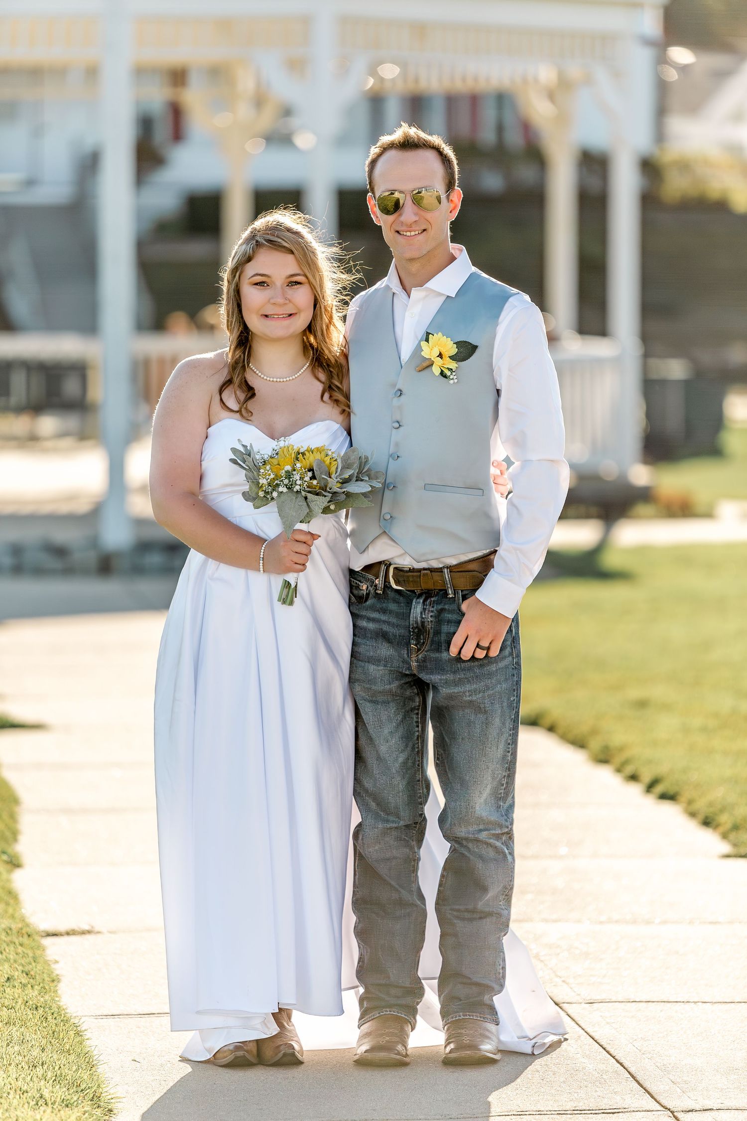 A couple in wedding attire poses outdoors, with the bride in a white dress holding a bouquet and groom in vest and jeans.