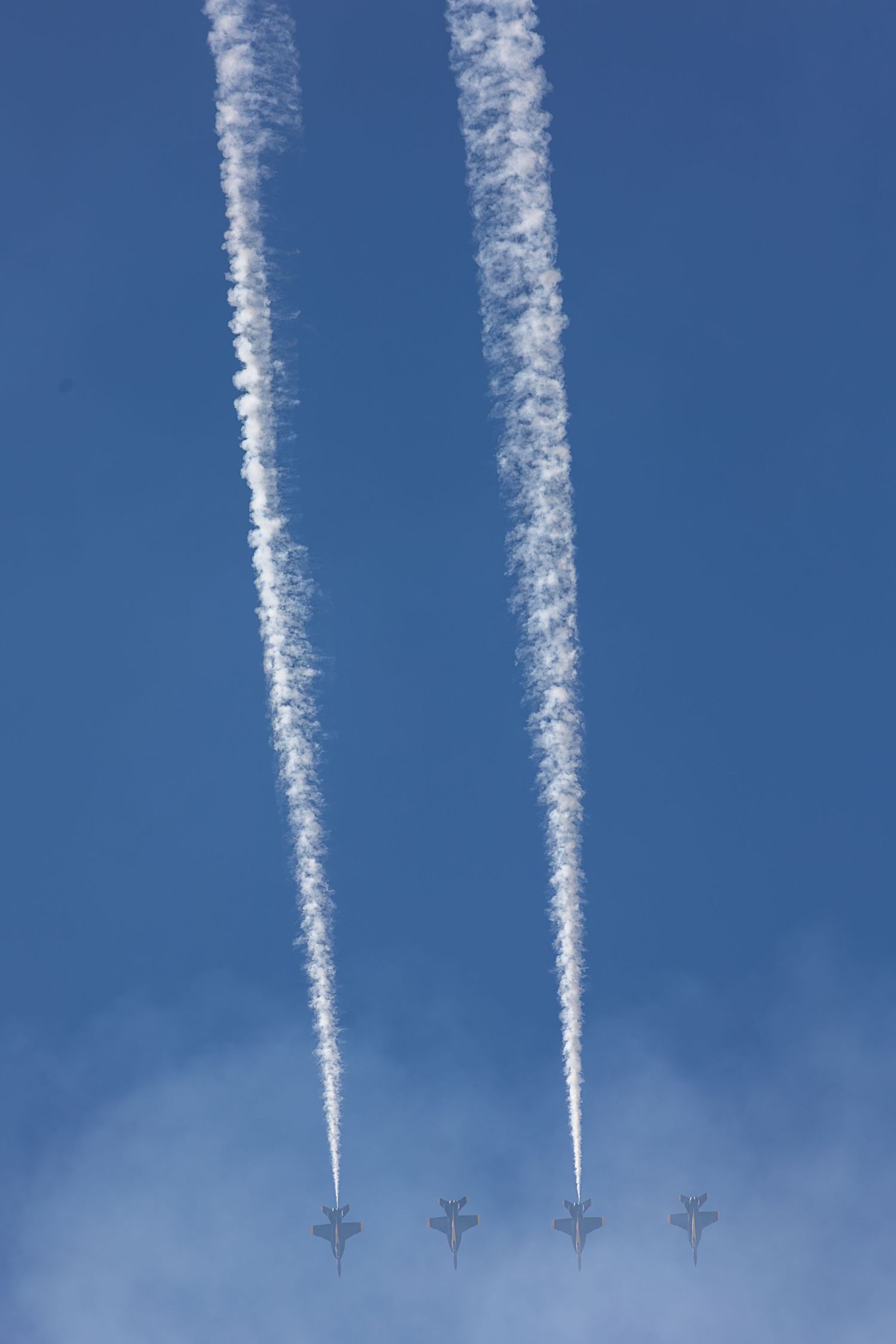 Contrails from multiple airplanes streak across a bright blue sky in a parallel formation pattern.