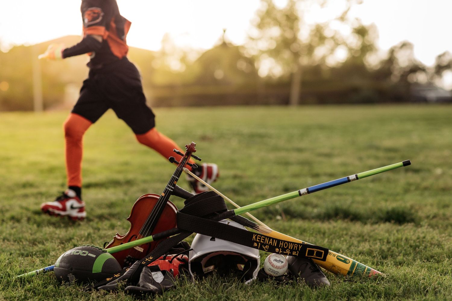 Sports equipment including lacrosse sticks, helmet, and a violin on grass with a player in background at sunset.