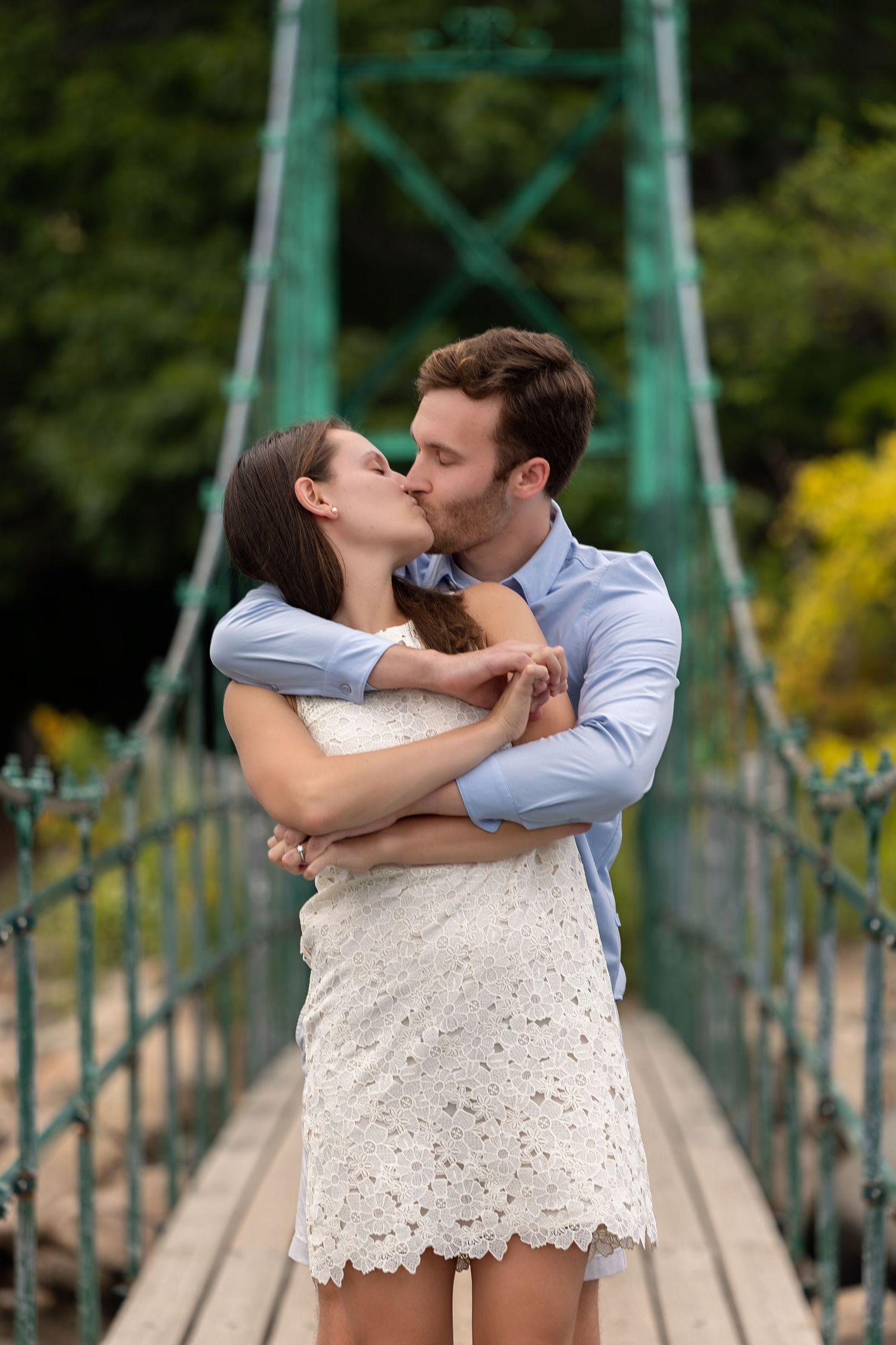 Couple sharing a romantic kiss on a suspension bridge, woman in white lace dress embraced by man in blue shirt.