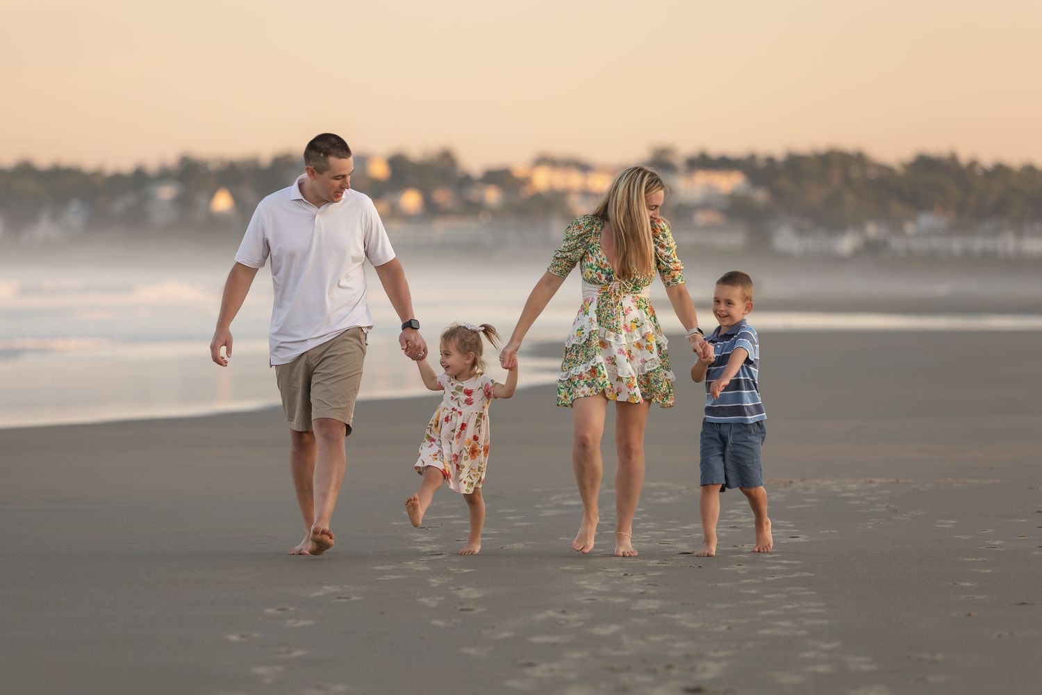 Family of four walking hand-in-hand on a sandy beach at sunset, with children in colorful outfits.