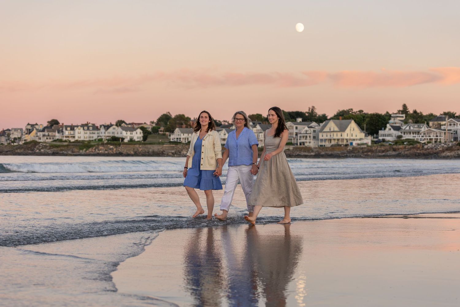 Three women walk barefoot on a beach at sunset with a full moon rising over coastal homes in the background.