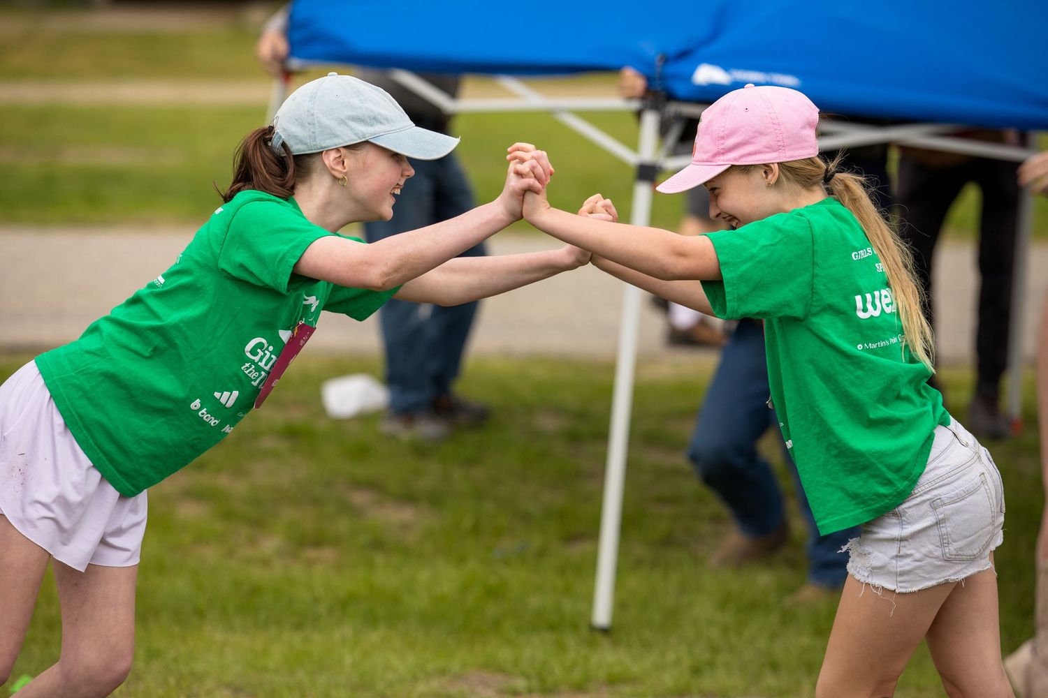 Two girls in green t-shirts playfully clasping hands outdoors at a summer event.