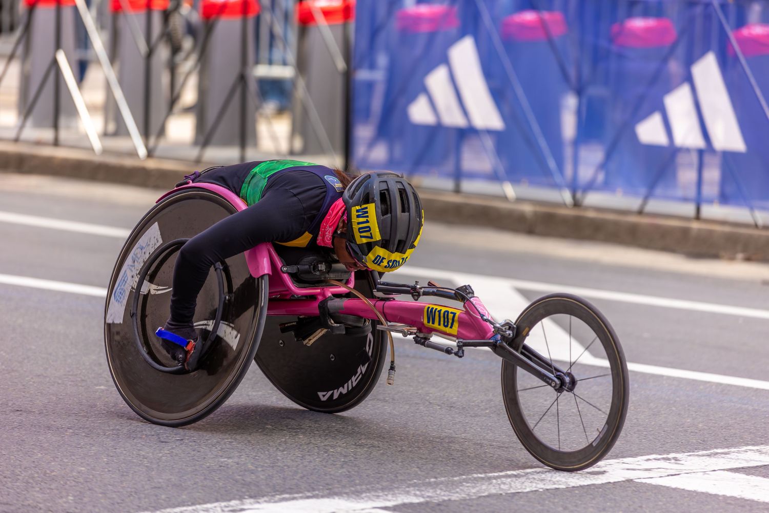 Paralympic athlete competing in a wheelchair race, leaning forward in a pink racing chair on a city marathon course.