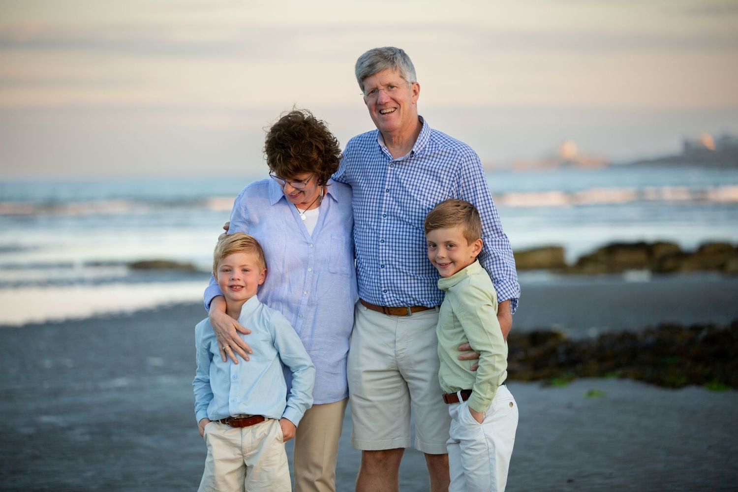 Family of four smiling on a beach at sunset, two adults and two young boys in coordinating light blue and khaki outfits.