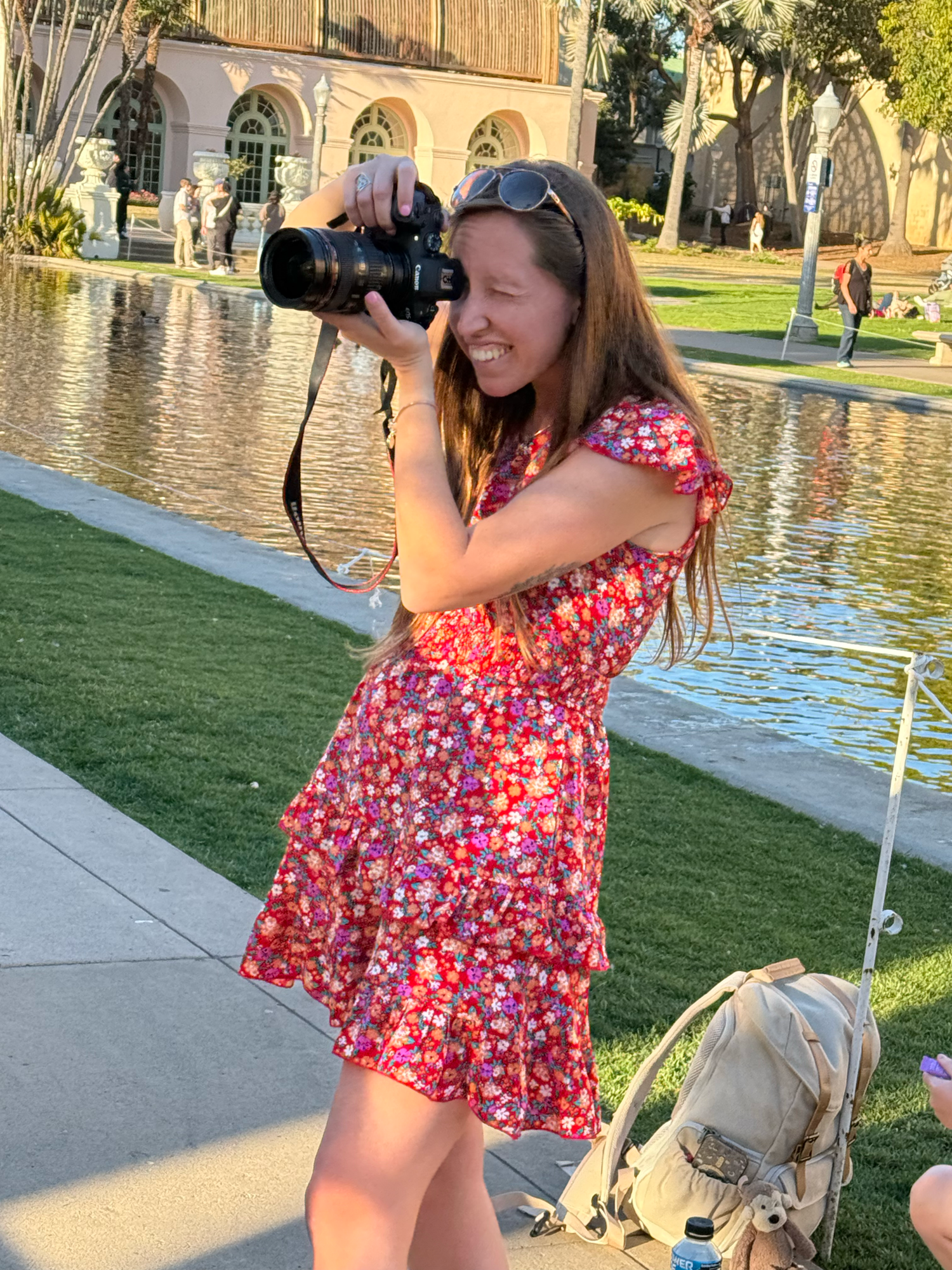 Photographer in a floral dress takes pictures by a reflecting pool on a sunny day at an ornate building.