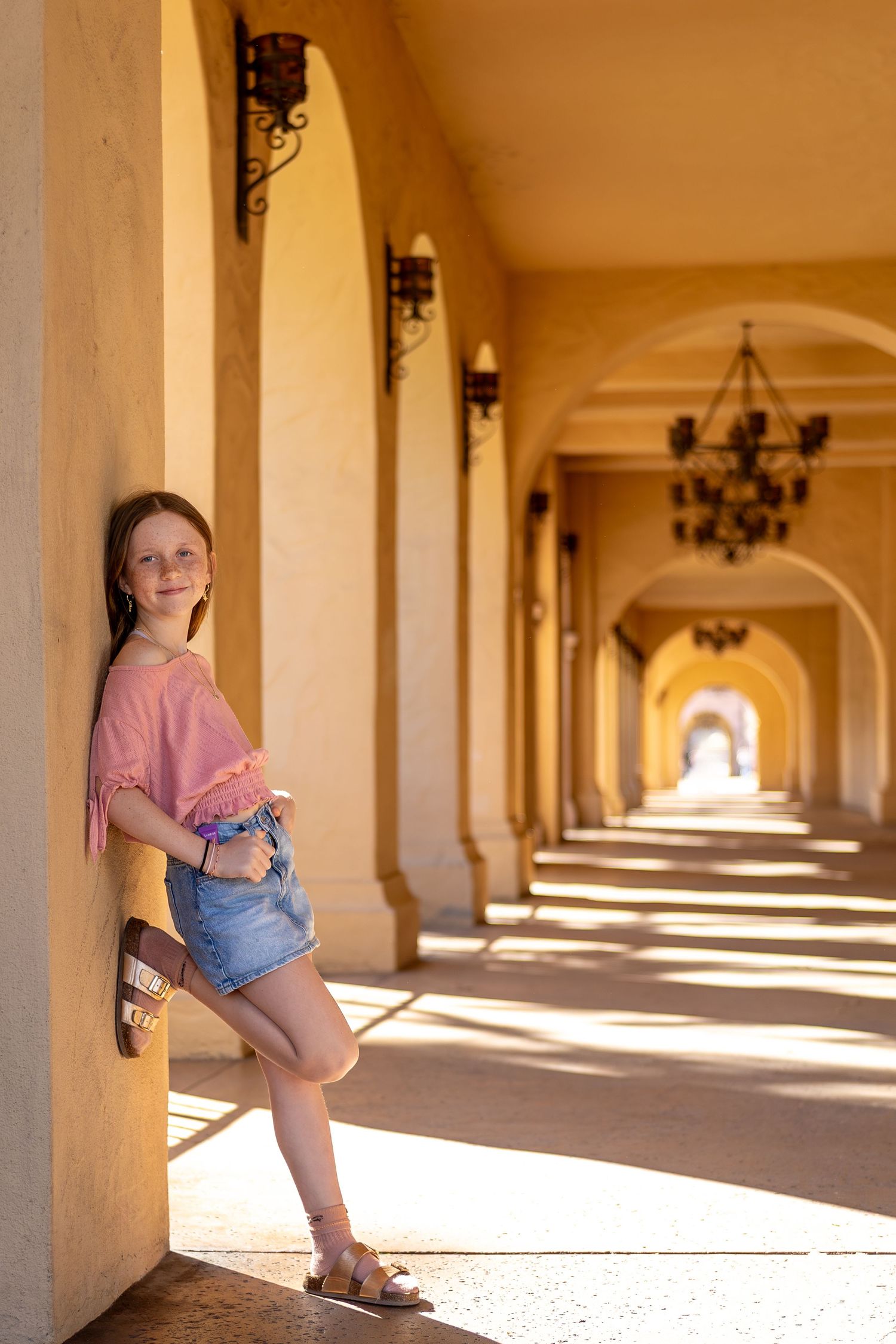 Young person posing in denim shorts against a sunlit colonnade with arched architecture.