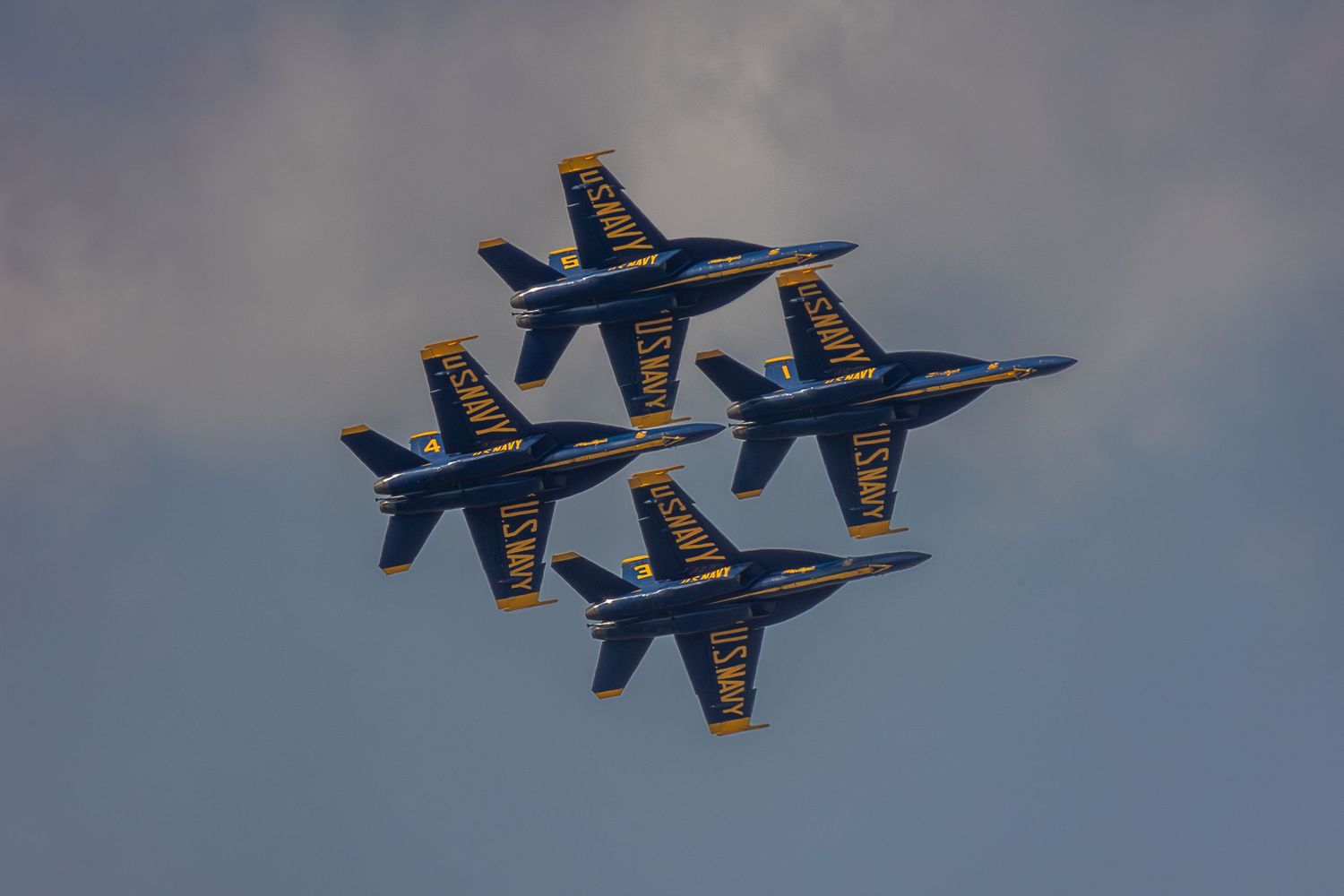 Blue and gold US Navy Blue Angels F/A-18 Hornets perform precision formation flying maneuvers against a cloudy sky.