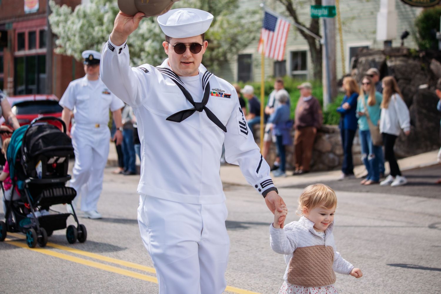 Navy sailor in white uniform waves while walking with toddler in a patriotic parade with American flag visible.