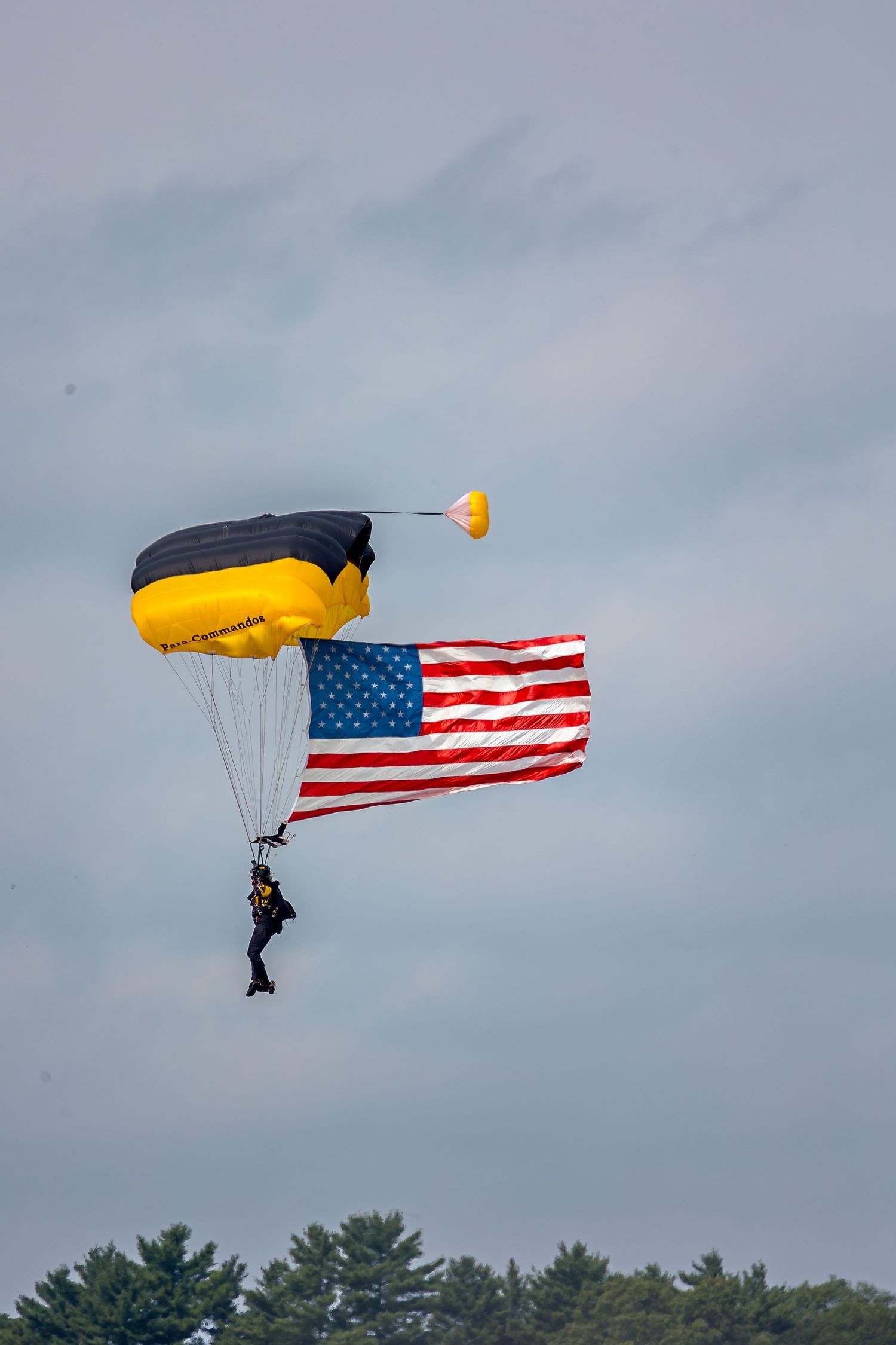 Skydiver with black and yellow parachute carrying a large American flag descends through cloudy skies at an airshow.