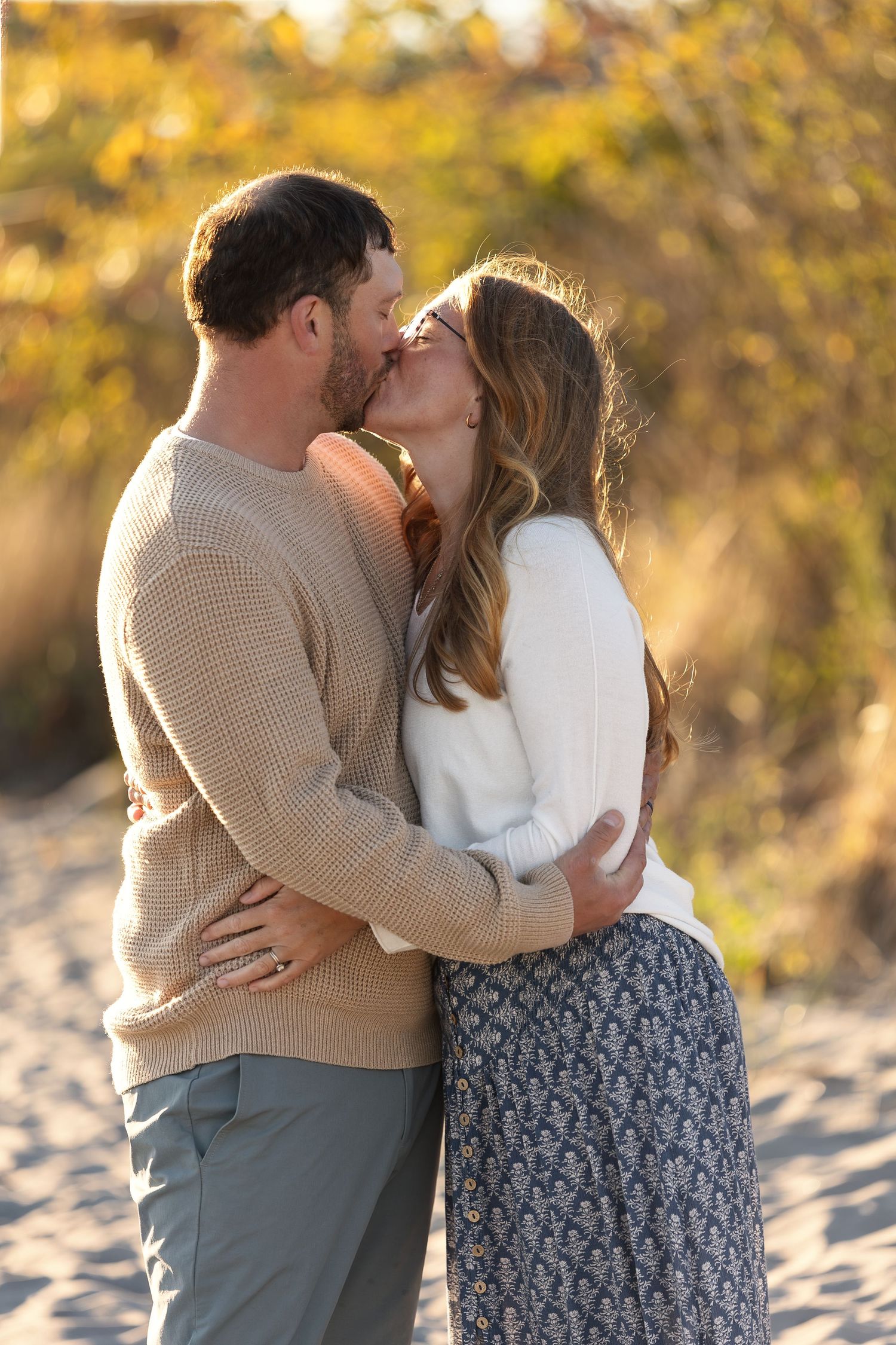 Couple sharing a romantic kiss on a sandy beach, embracing in cozy fall sweaters with golden autumn foliage backdrop.