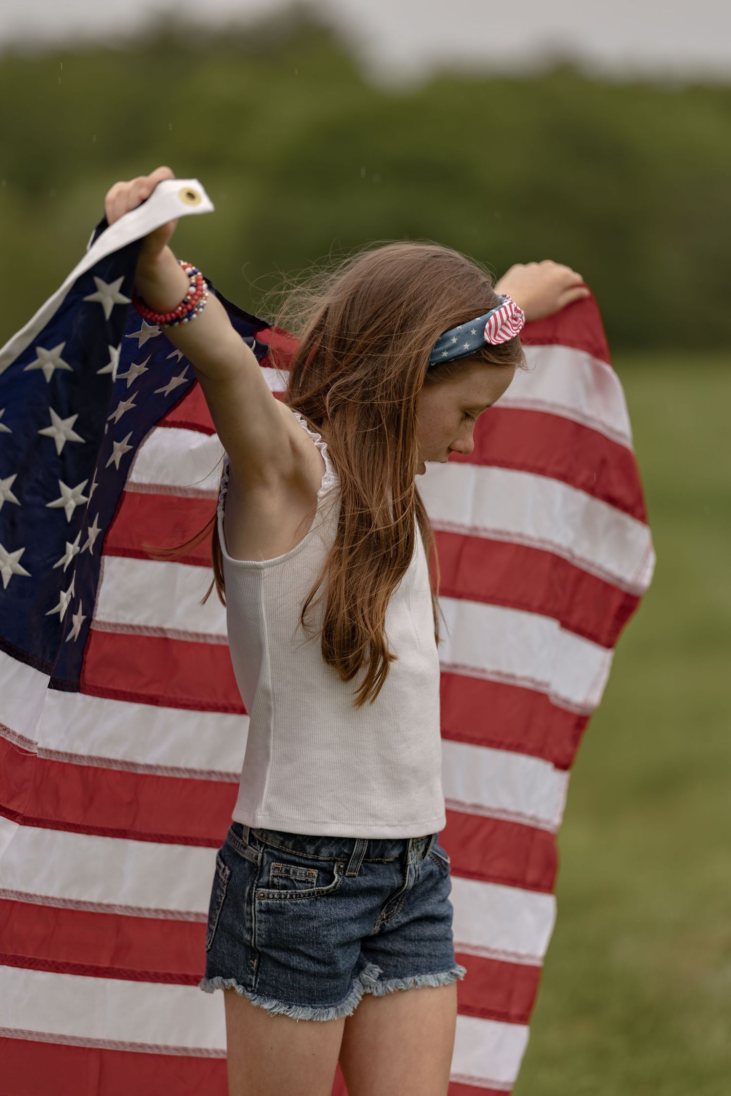 Young girl holding American flag outdoors in field, wearing denim shorts and white top with patriotic accessories.