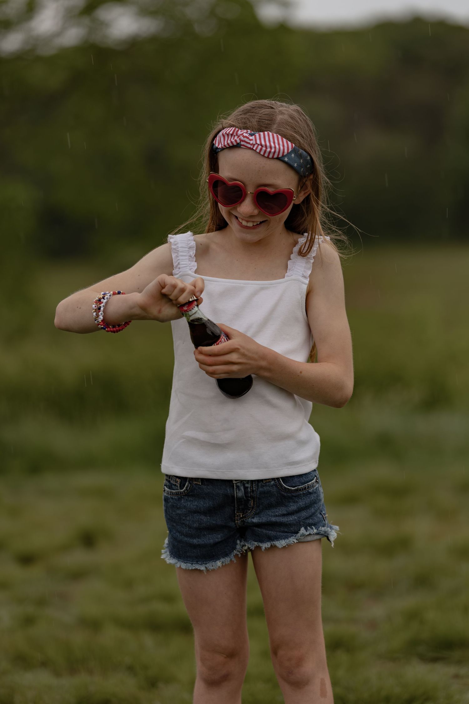 Young girl in heart sunglasses and denim shorts opening a Coca-Cola bottle outdoors in a green field.