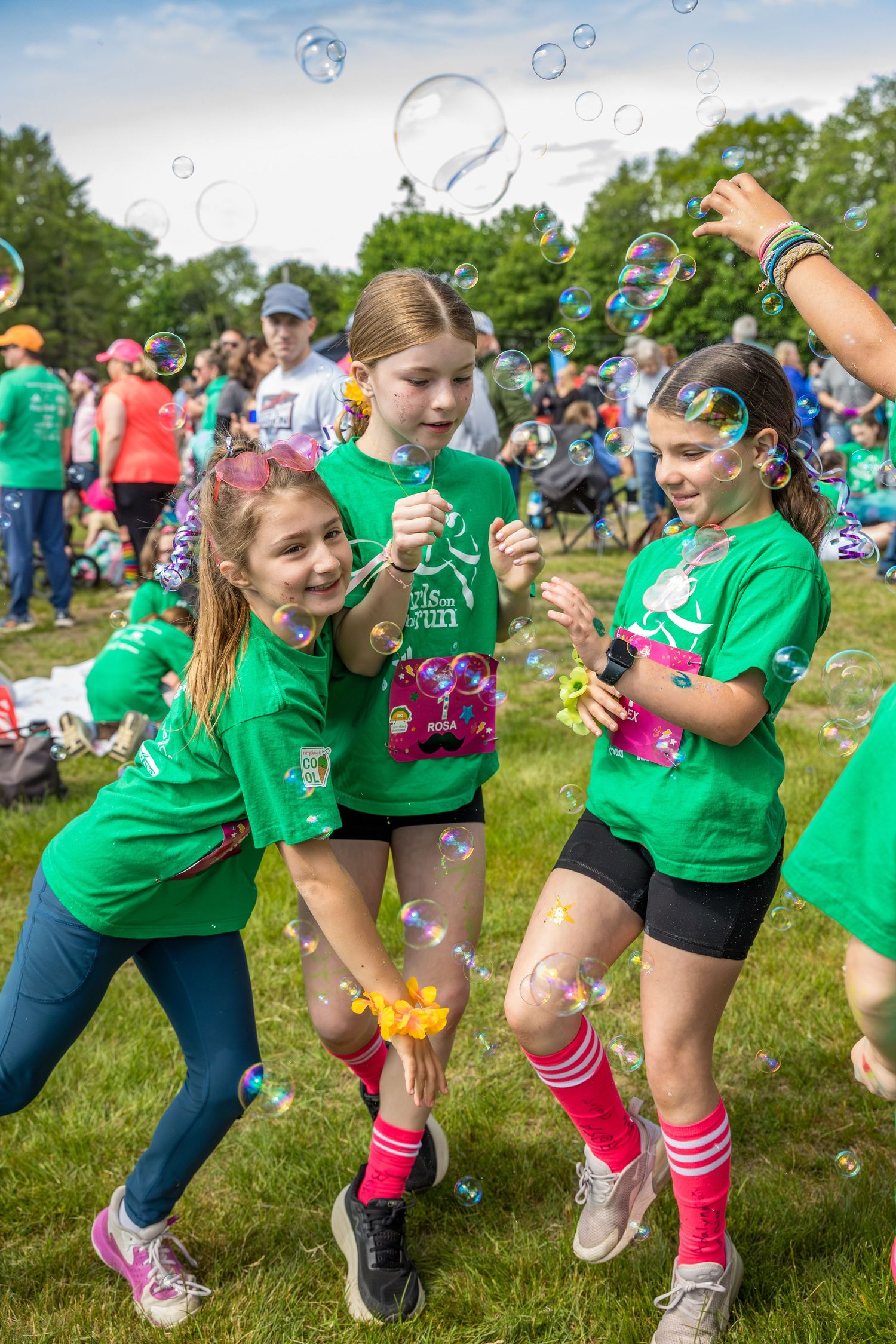 Three girls in green t-shirts laugh and play with bubbles outdoors at a fun run event on a grassy field.