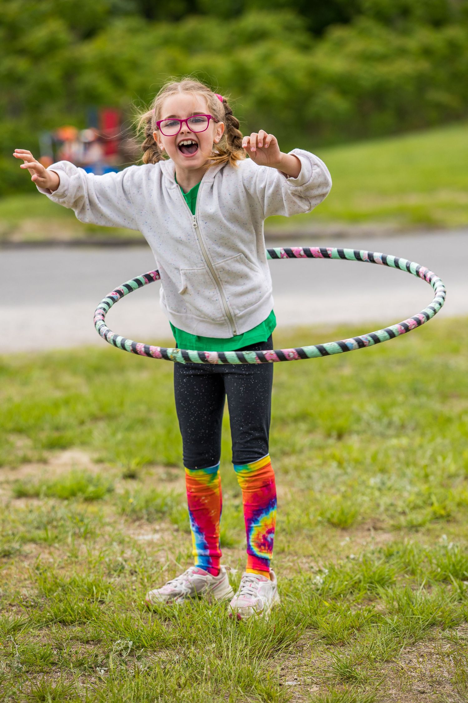A happy young girl with glasses hula hooping outdoors, wearing tie-dye socks and a gray hoodie in a grassy park.