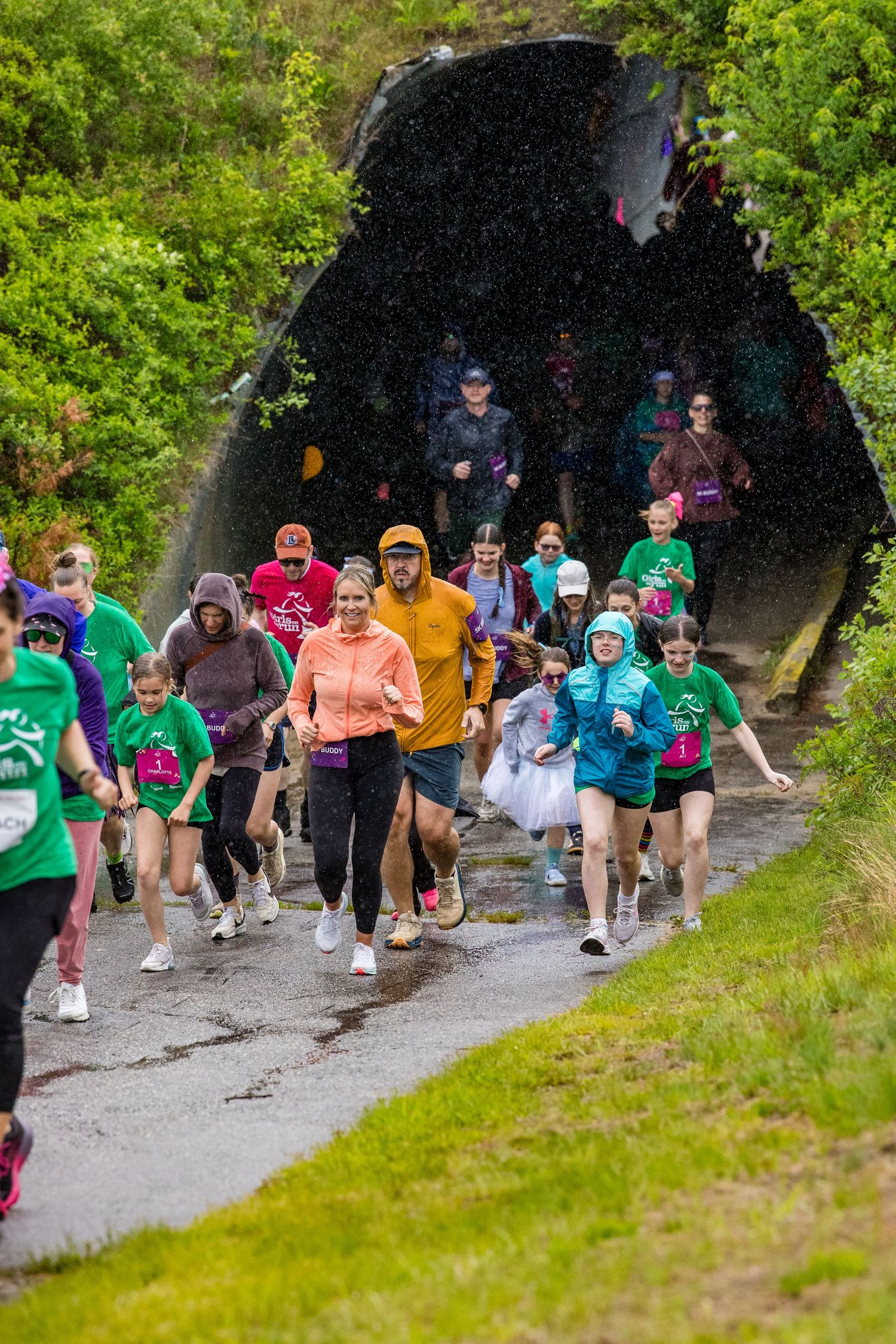 Runners of all ages emerge from a tunnel on a rainy day during a community fun run event.